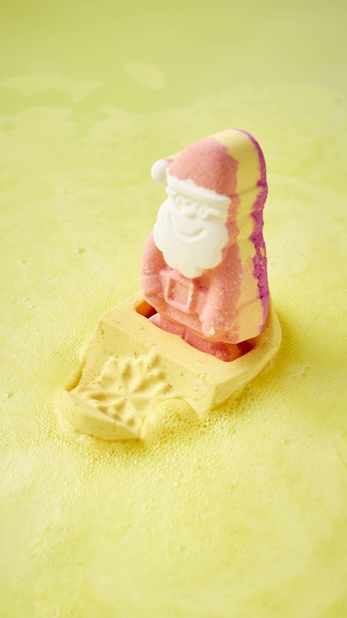 The image shows the festive, Magical Santa Sleigh, two-part bath bomb sitting upright, balanced in the bath water as it begins to give off bright, sunshine-yellow foam. 