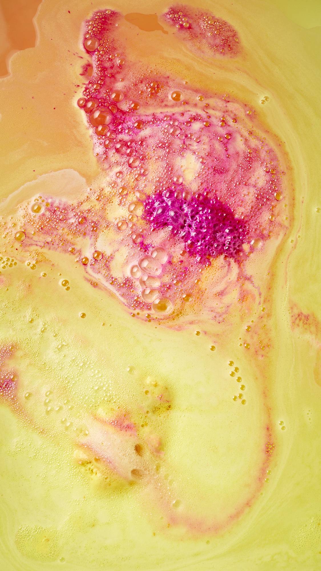 The image shows an aerial view of the the bath water after using the festive Magical Santas Sleigh, two-part bath bomb. There is a bubbly blanket of contrasting yellow, pink and red foam covering the surface. 