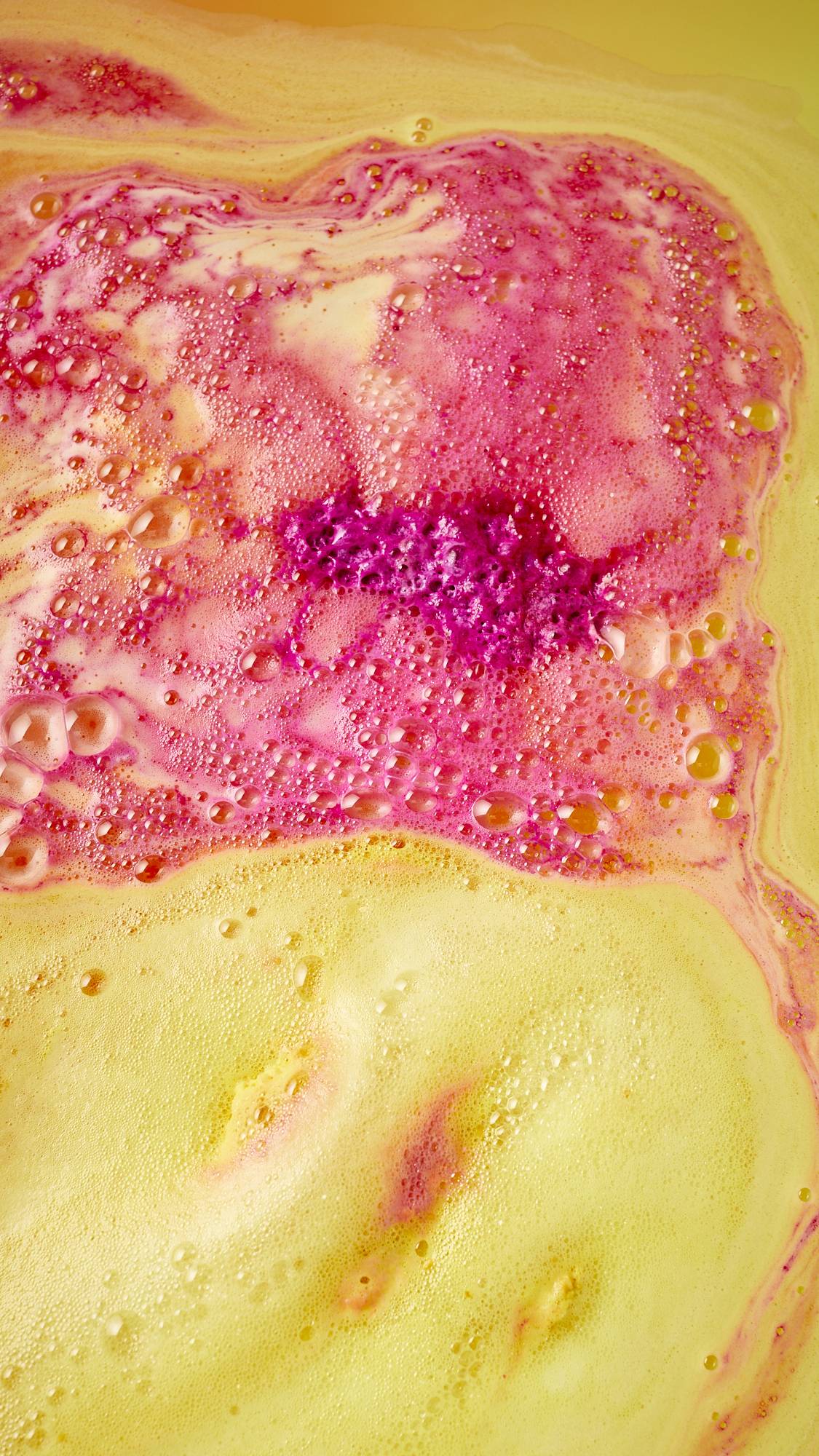 The image shows an aerial view of the the bath water after using the festive Magical Santas Sleigh, two-part bath bomb. There is a bubbly blanket of contrasting yellow, pink and red foam covering the surface. 