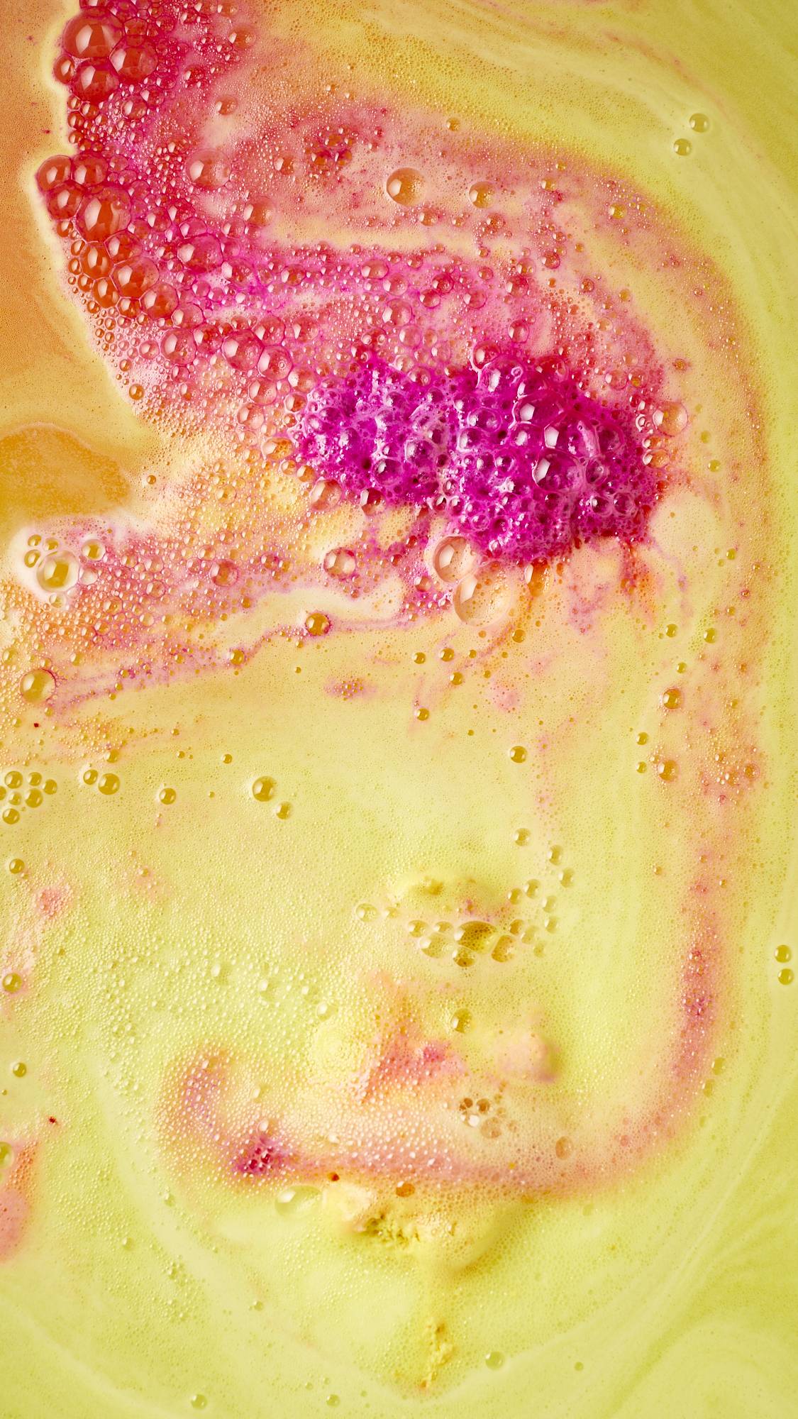 The image shows an aerial view of the the bath water after using the festive Magical Santas Sleigh, two-part bath bomb. There is a bubbly blanket of contrasting yellow, pink and red foam covering the surface. 