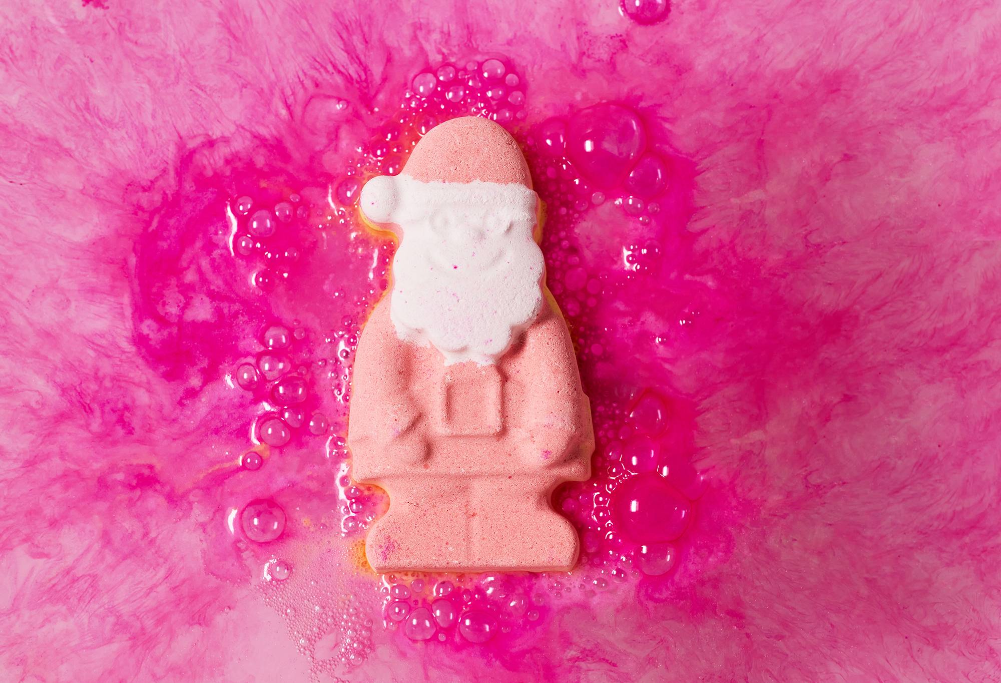 Image shows Magical Santa bath bomb laying on top of bath water. Vivid, hot-pink swirls branch out from behind.