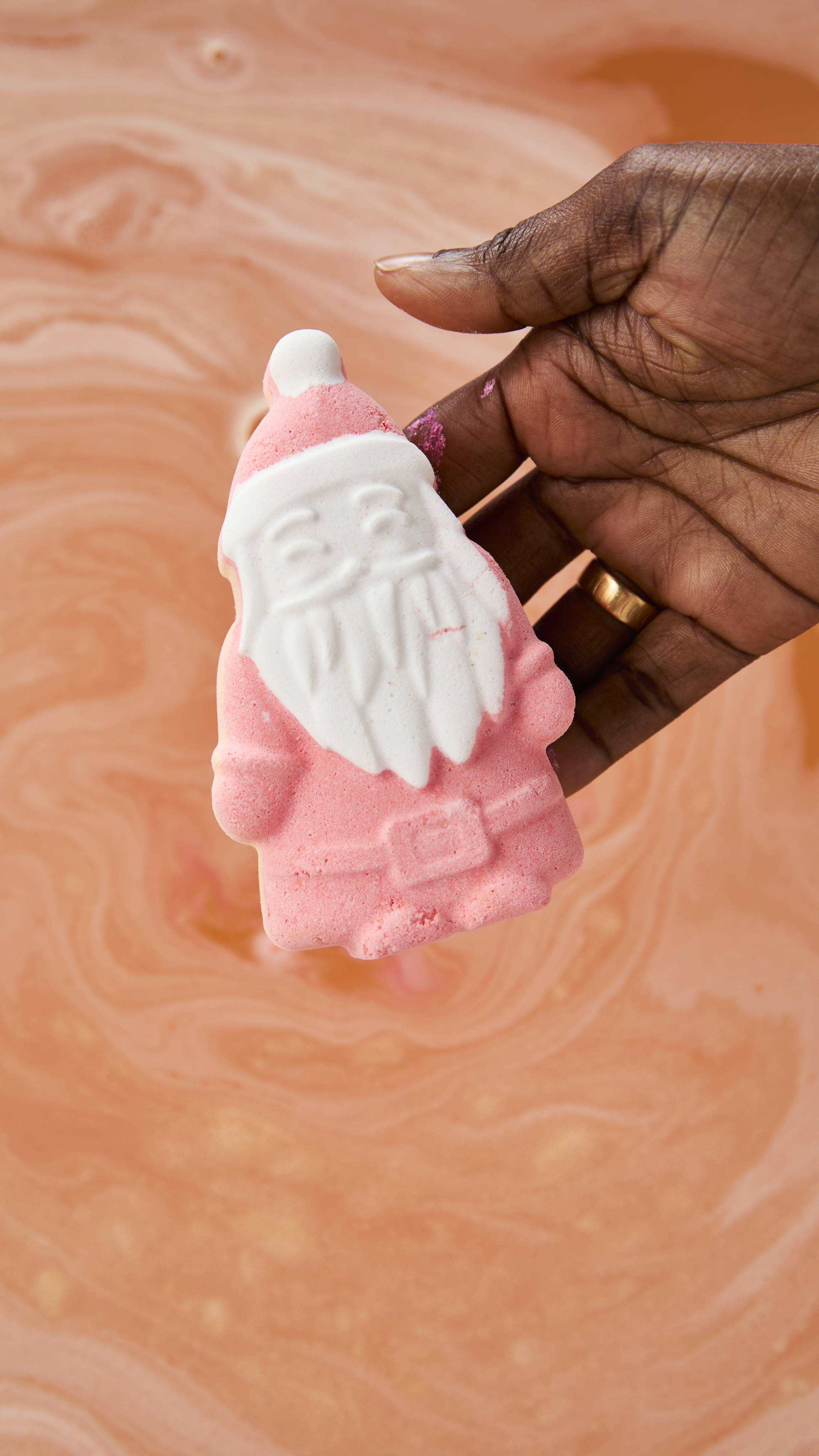 A hand holds a pink Lush "Magical Santa" bath bomb with a white beard and hat trim over swirling, peachy-pink bathwater.