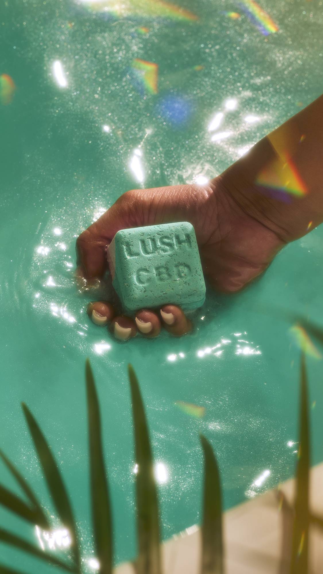 A close-up image of the model's hand holding the Magik bath bomb just under the water creating ripples of shimmering teal water. Some plant leaves are just visible and there are flecks of rainbow reflections. 
