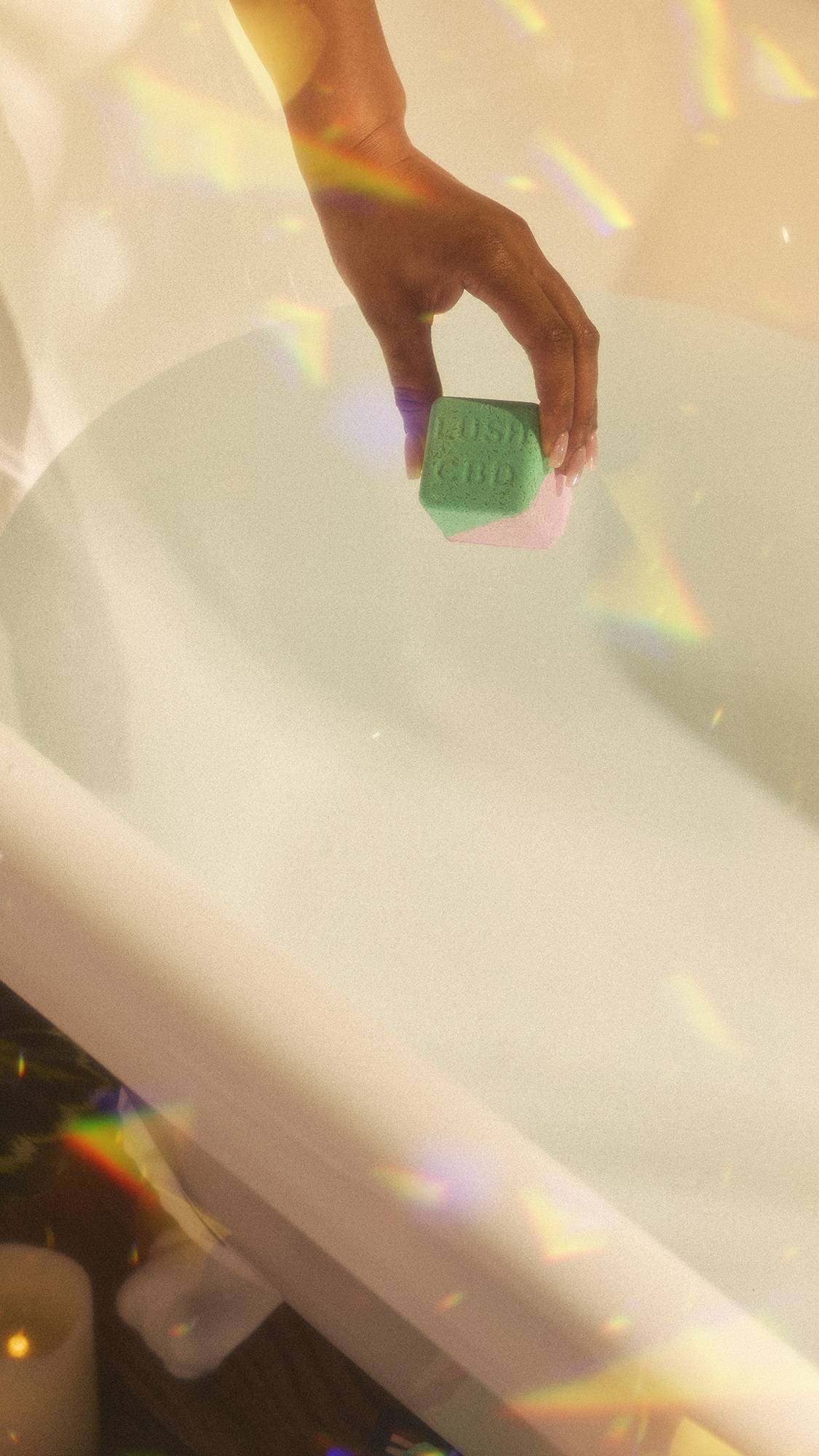 A close-up of the model's hand holding the Magik CBD Epson Salt bath bomb over the still bath water in the tub. The image has flecks of rainbow reflections on a warm, sepia tone.