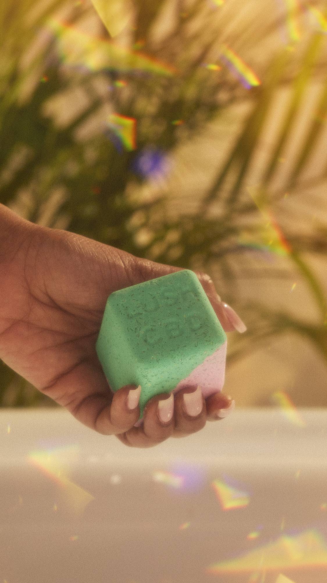 A close-up of the model's hand holding the Magik CBD Epson Salt bath bomb focusing on the vibrant pink and green. The image has flecks of rainbow reflections on a warm, sepia tone.