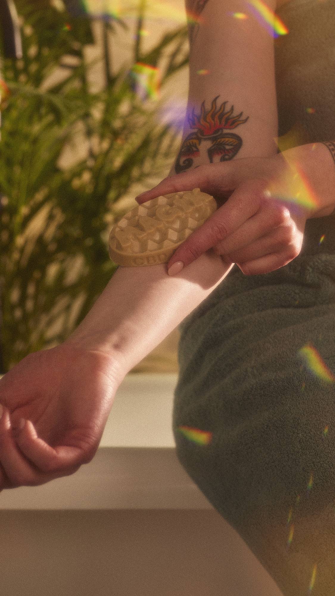 A close-up image of the model holding the Magik massage bar and smoothing it over their forearm. The image has flecks of rainbow reflections on a warm, sepia tone.