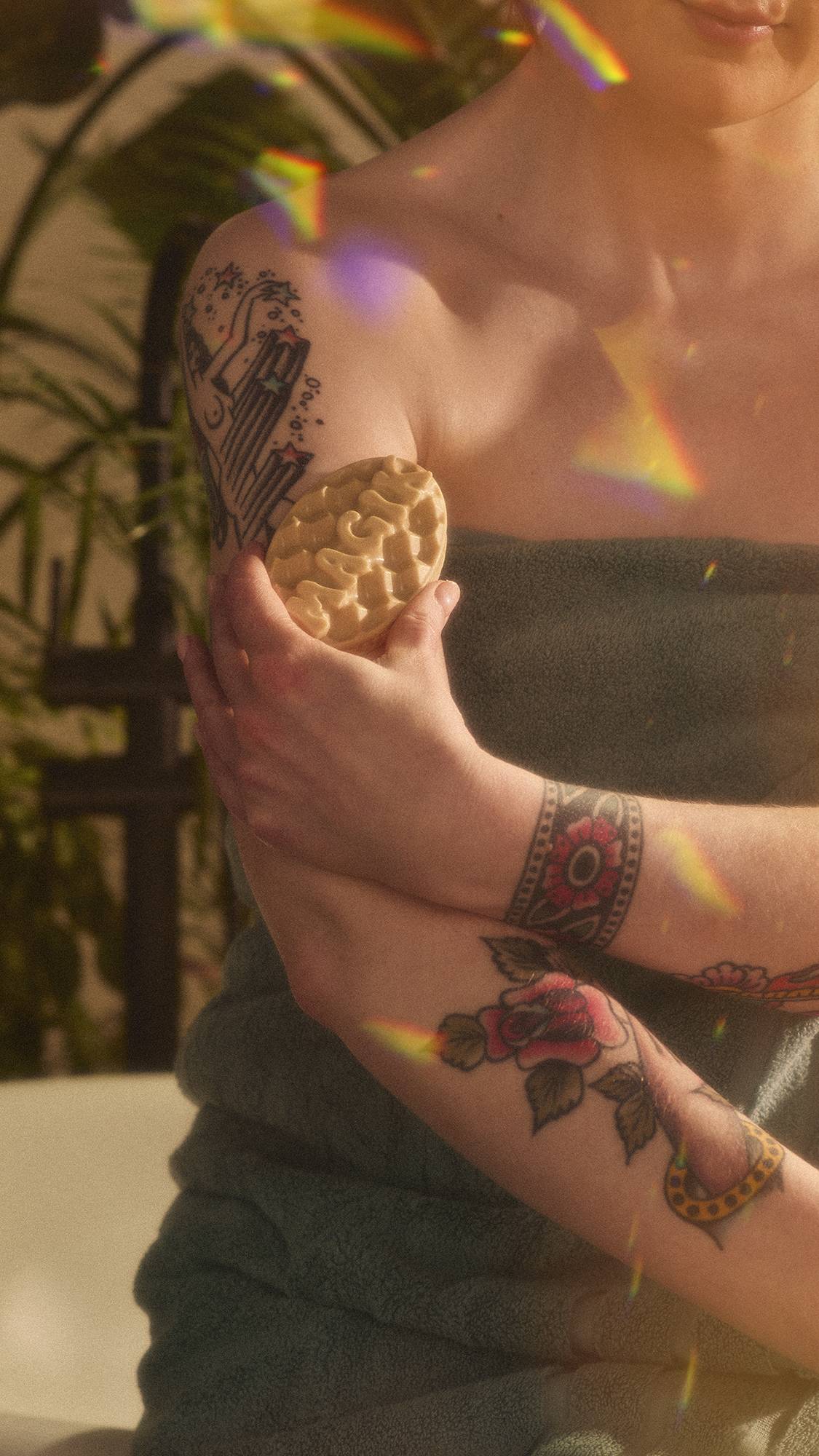 A close-up of the model's torso as they are wrapped in a sage green towel sitting on the edge of the bathtub using the Magik massage bar over their upper arm. The image has flecks of rainbow reflections on a warm, sepia tone.