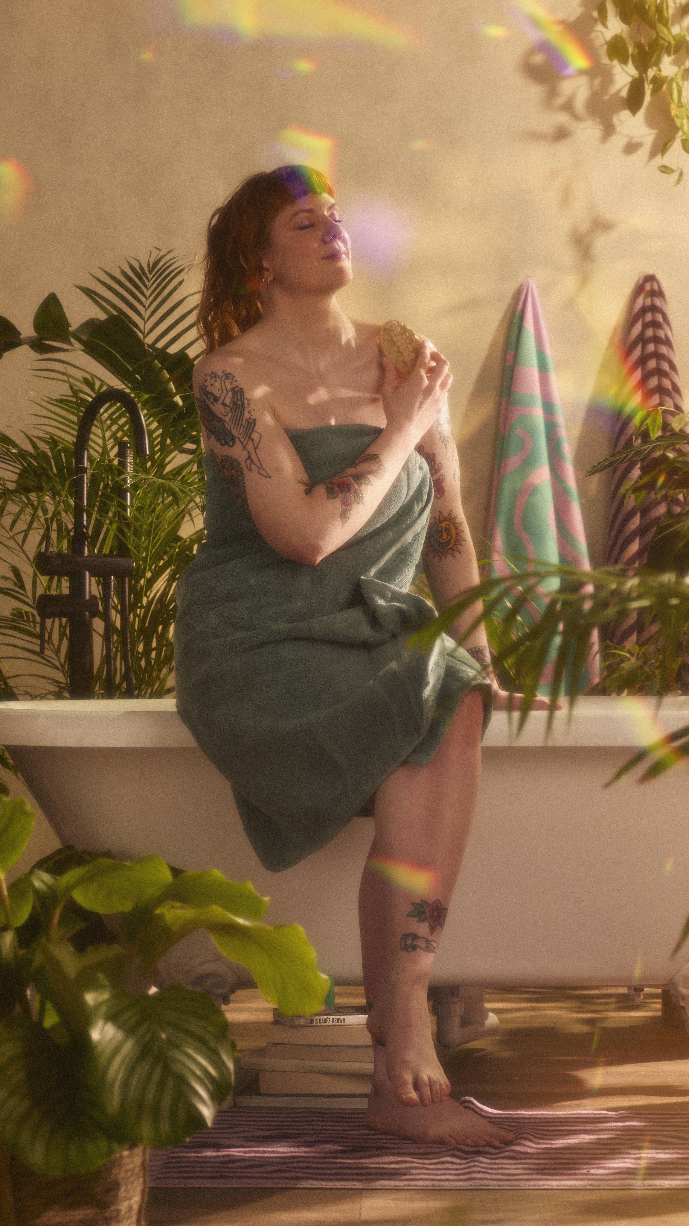 The model is wrapped in a sage green towel while sitting on the edge of a roll-top bathtub using the Magik massage bar over their chest. The image has flecks of rainbow reflections on a warm, sepia tone.