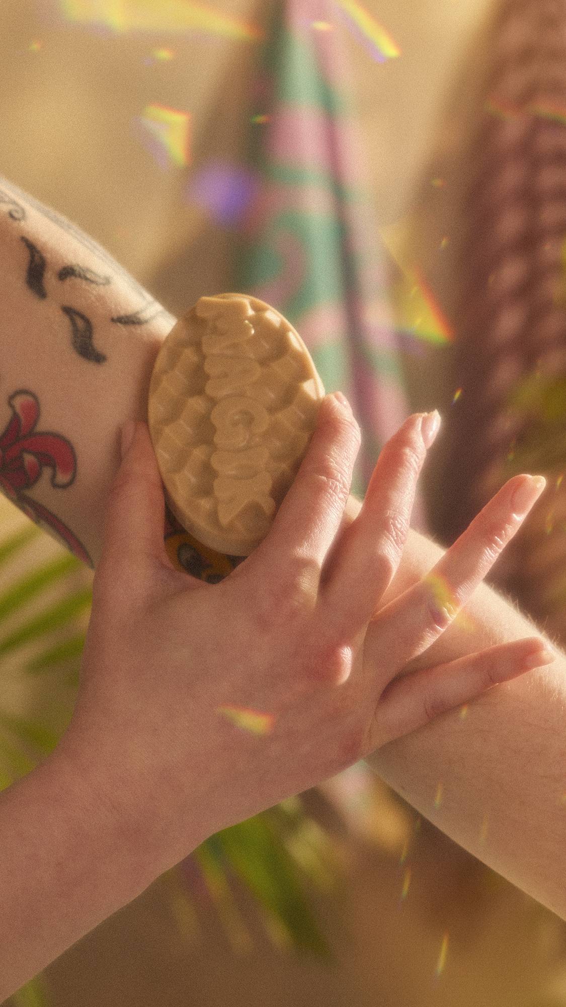 A close-up image of the model holding the Magik massage bar and smoothing it over their forearm. The image has flecks of rainbow reflections on a warm, sepia tone.