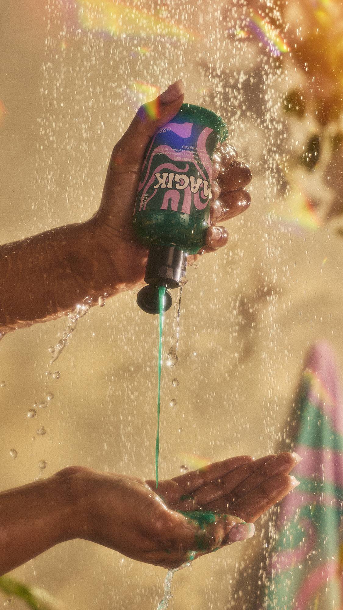 A close-up of the model's hands under running shower water as they squeeze the thick, shimmering green shower gel from the product bottle into their hand. The image has flecks of rainbow reflections on a warm, sepia tone. 