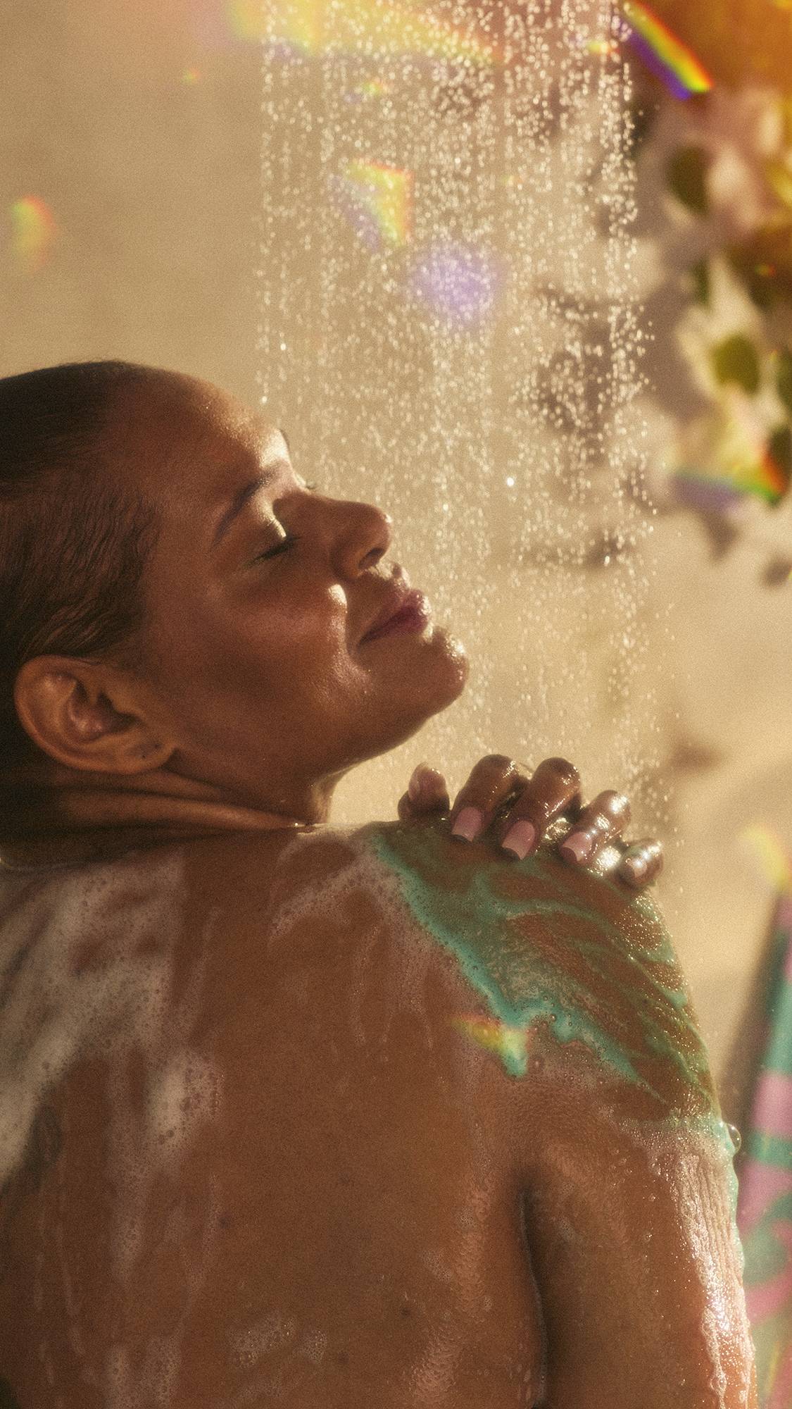 The model is under the running shower water facing away as they lather the Magik shower gel over their shoulder. The image has flecks of rainbow reflections on a warm, sepia tone.