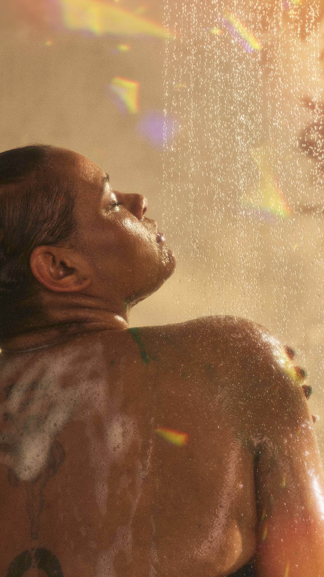 The model is under the running shower water facing away as they lather the Magik shower gel over their shoulder. The image has flecks of rainbow reflections on a warm, sepia tone.