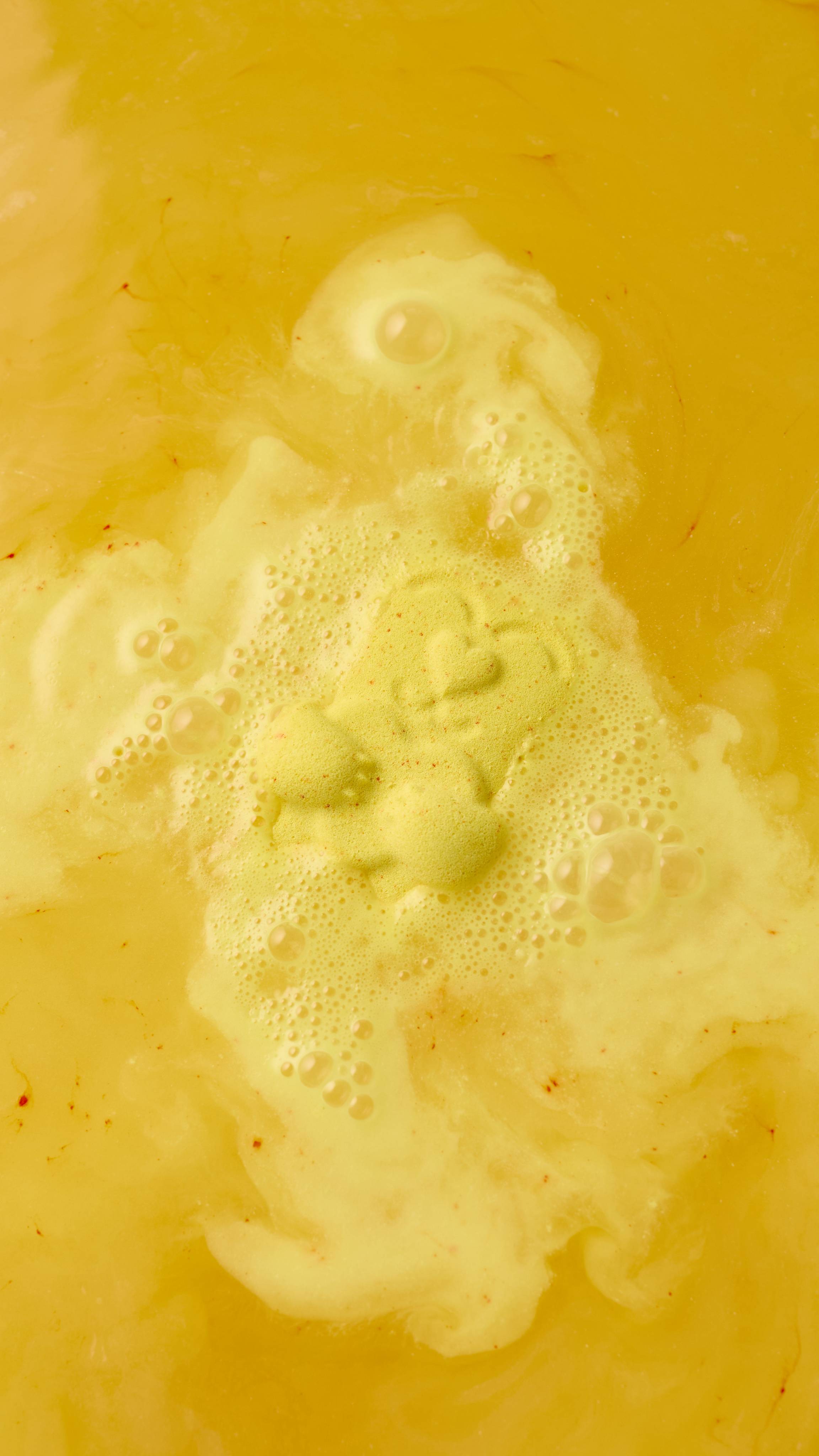 A yellow "Mama Bear" bath bomb dissolves in golden bathwater, creating a frothy layer of creamy white bubbles. The bear shape, featuring a small heart on its chest, is visible amidst the fizzing foam as it melts into the warm, yellow-orange water.