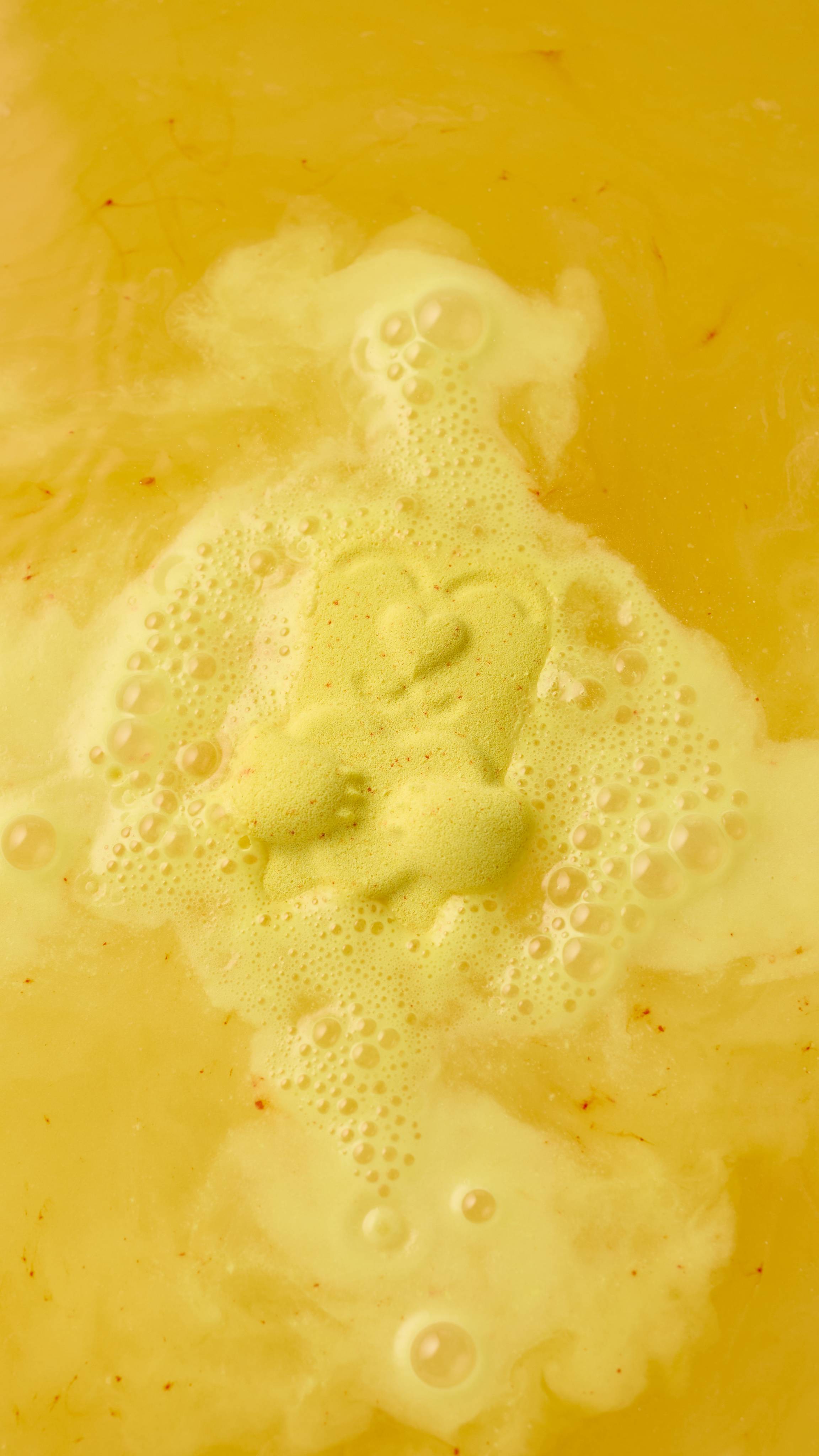 A dissolving yellow "Mama Bear" bath bomb fizzes in water, releasing a creamy foam. The bath bomb shape is still faintly visible in the center of the froth, surrounded by golden-yellow water.