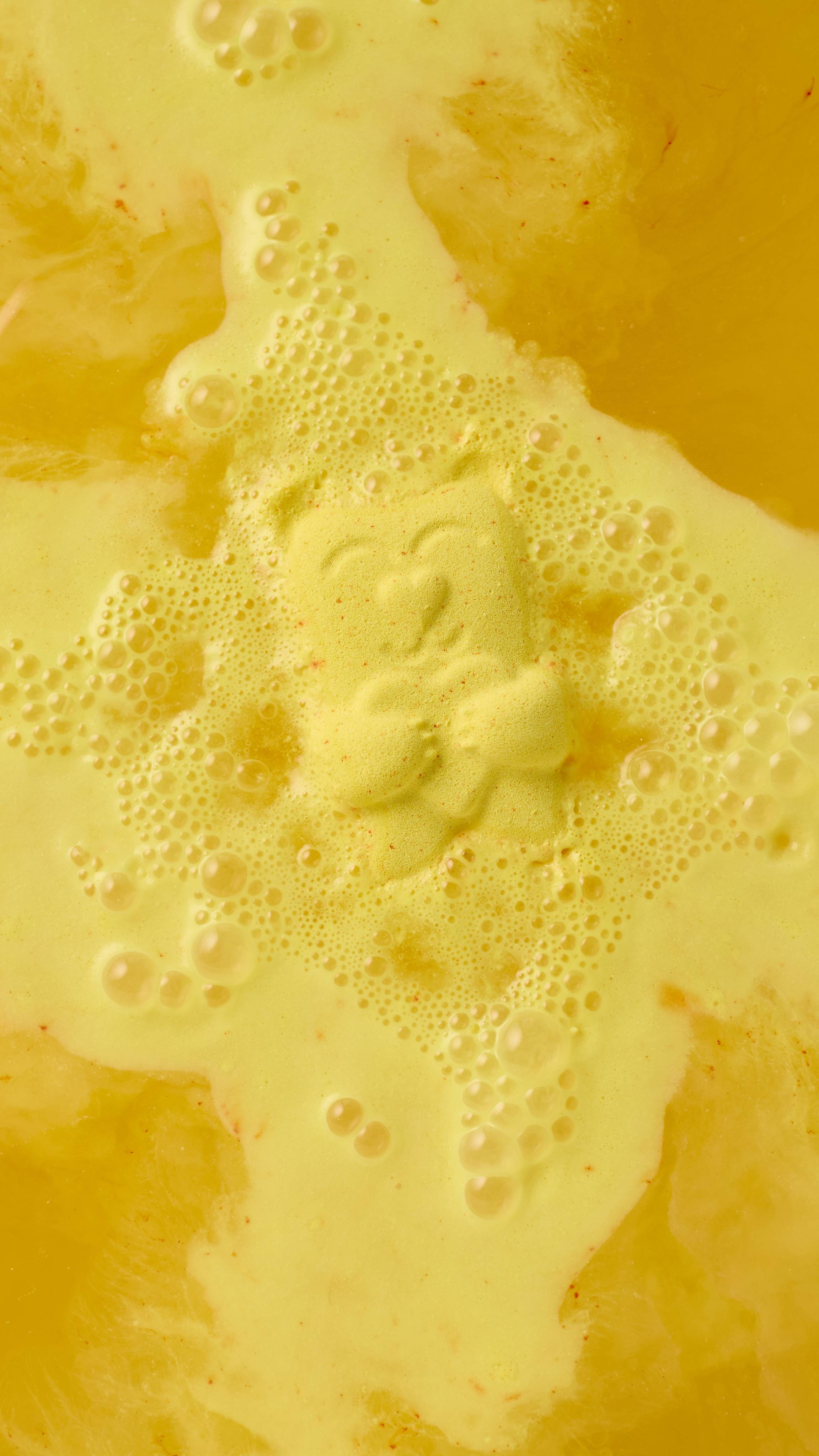 A yellow "Mama Bear" bath bomb floats in golden bathwater, dissolving into a creamy froth of bubbles. The bear's smiling face and the heart on its chest are faintly visible as the fizzer melts away, turning the water a rich yellow-orange hue.