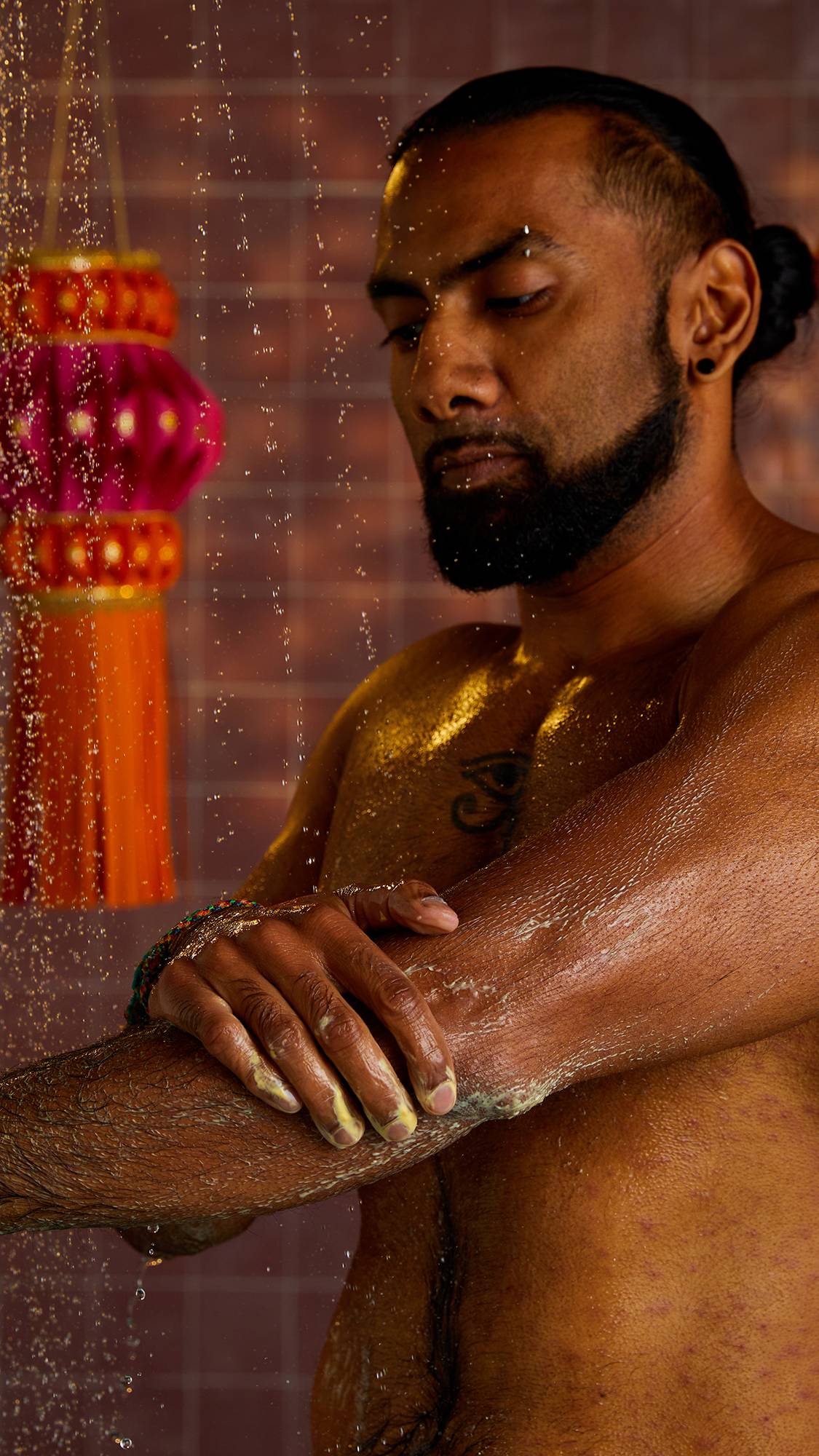 A man with a beard and tattoos stands in a shower, with his eyes closed, as he rubs his arm with a solid, yellow shower gel. In the background are decorative tassels and a tiled wall.