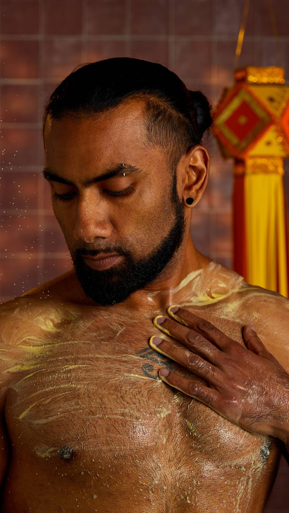 A man with a beard stands in the shower, looking down at his chest. He is lathering his body with a yellow shower gel. In the background are decorative tassels and a tiled wall.
