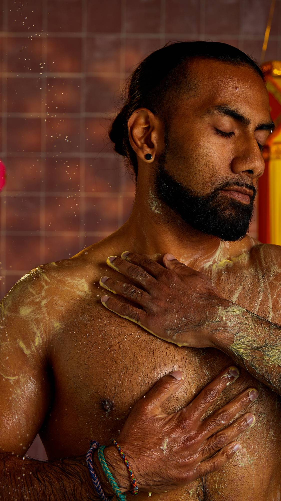 A man with a beard stands in the shower, with his eyes closed, and his hands on his chest. He is lathering his body with a yellow shower gel. In the background are decorative tassels and a tiled wall.