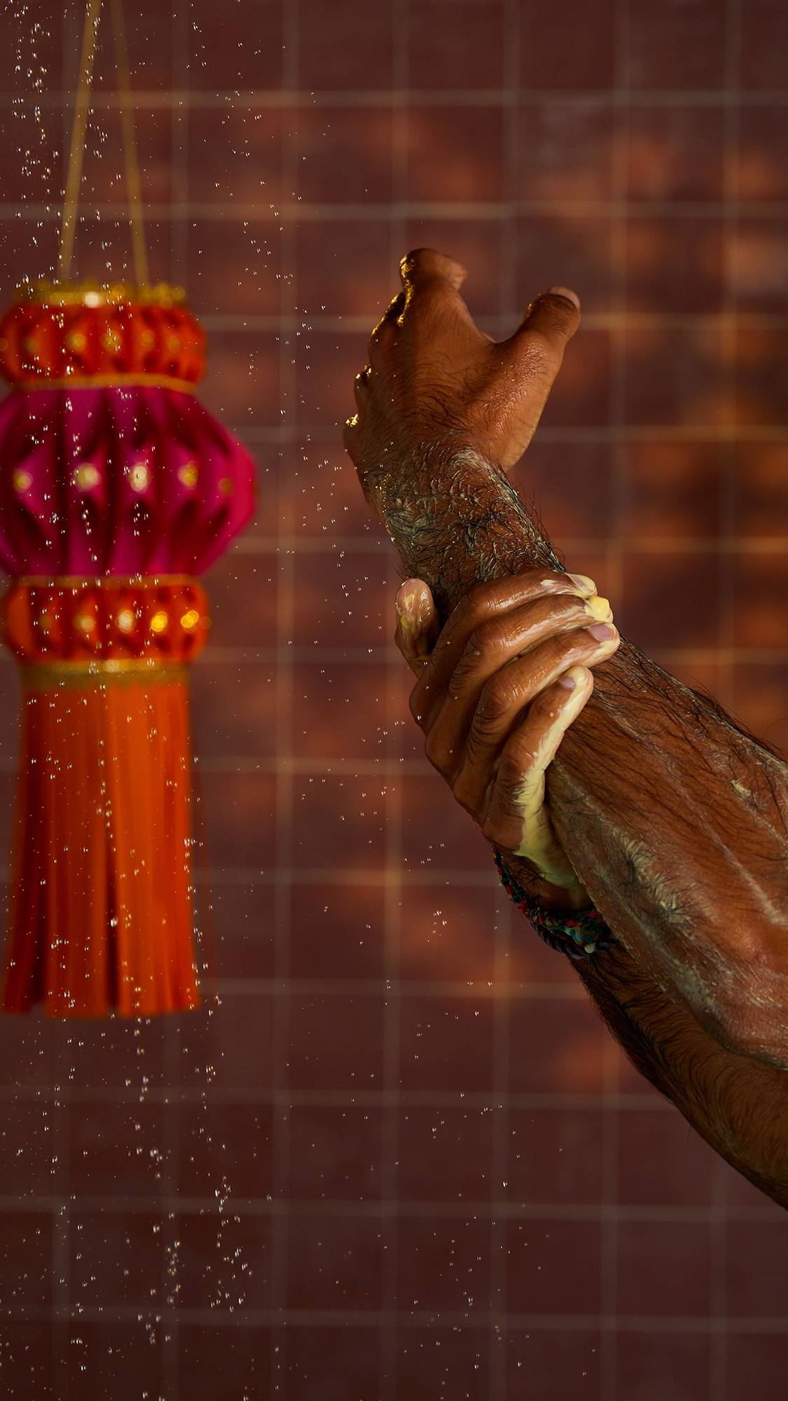 A close-up shot of a hand and arm as it lathers with shower gel in the shower. Water droplets are suspended in the air. In the background is a tiled wall and a red and orange decorative tassel.