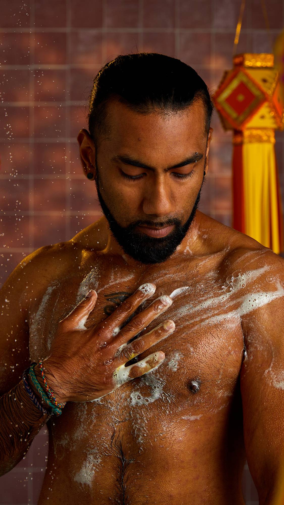 A person with a beard and tattoos stands in a shower, lathering his chest with white foam. His eyes are closed and a gentle spray of water falls around him, creating a refreshing scene.
