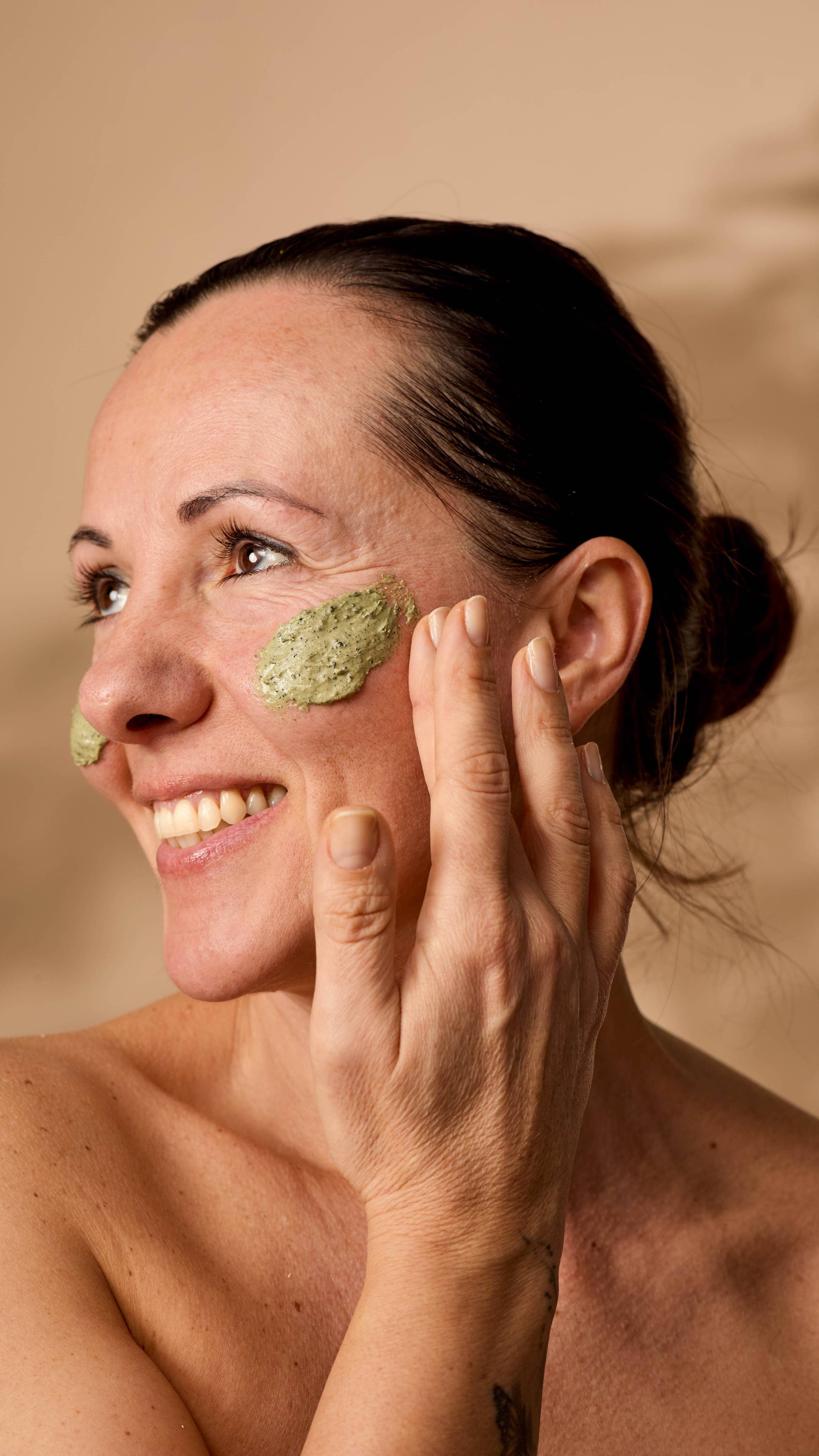 The model is smiling as they apply the green Matcha face mask to their other cheek under warm lighting.