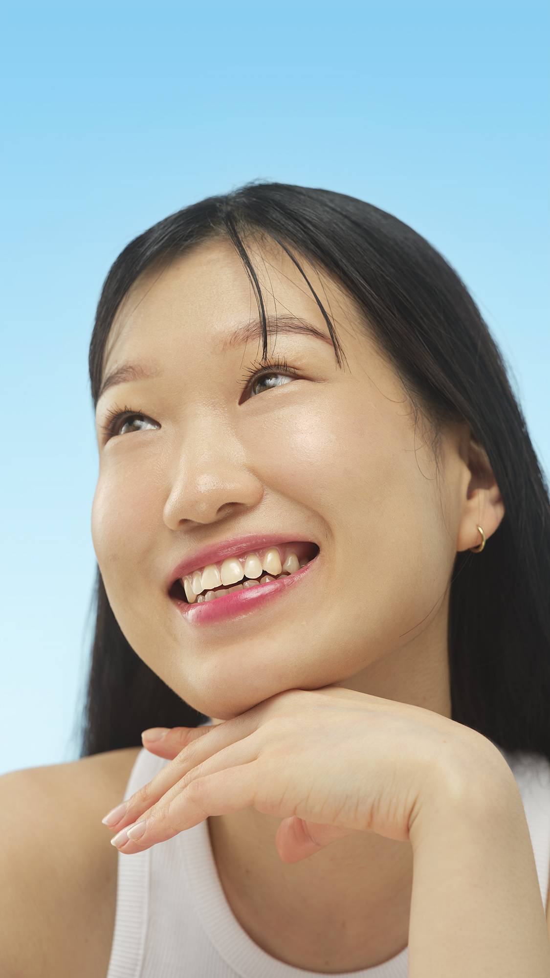A close-up vertical portrait of a smiling person with cool-toned skin and dark hair, looking up and left. Their chin rests on their hand, and their face has a subtle glow from the Medium Cool Beauty Balm, set against a bright blue background.