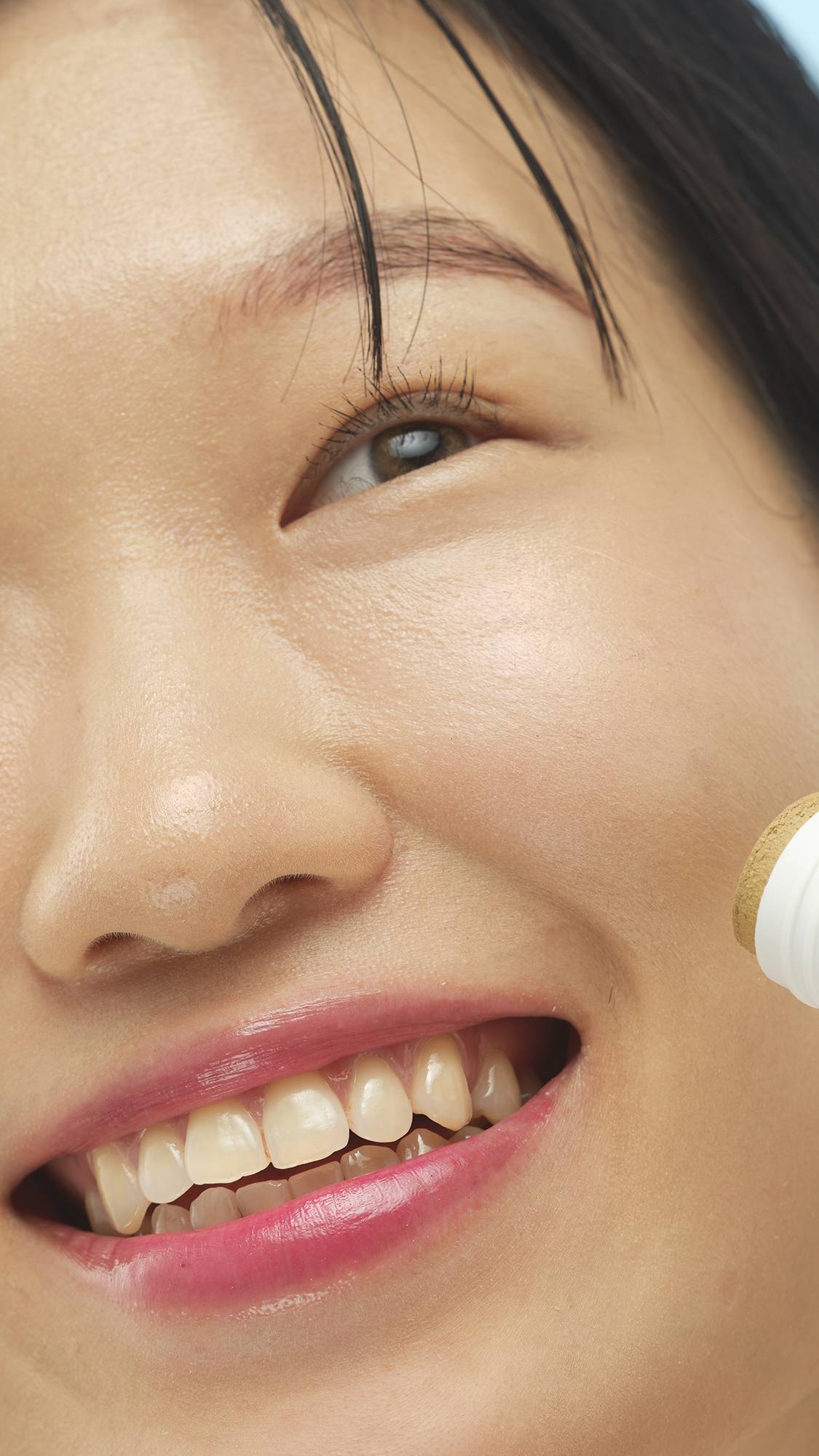 A close-up vertical shot of a person's smiling face, showing their eye and mouth. They have cool-toned, medium skin, and the stick of Medium Cool Beauty Balm is visible at the edge of their cheek.
