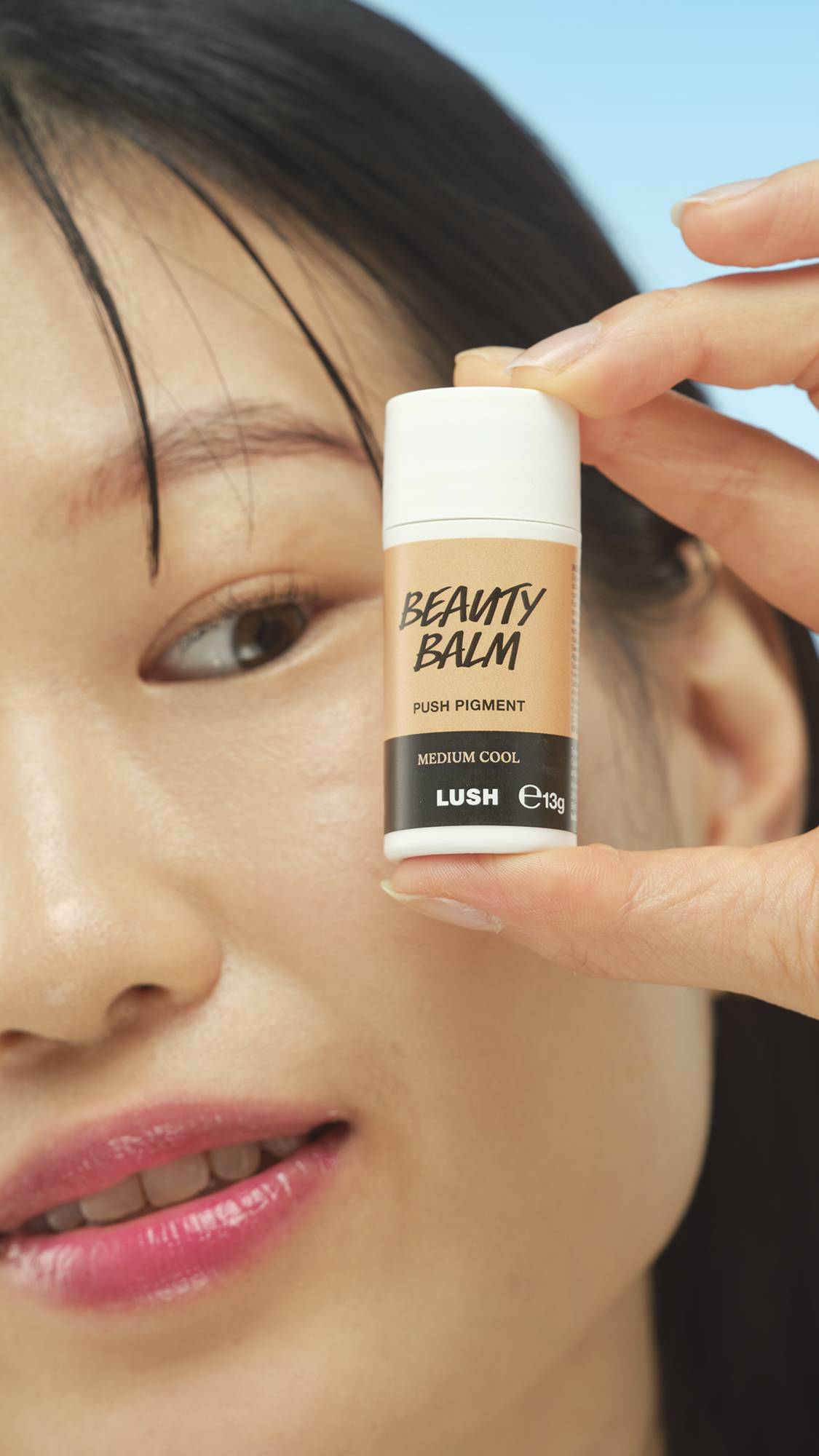 A close-up vertical shot of a person's smiling face, with a hand holding the white and beige stick of Medium Cool Beauty Balm right next to their cheek, showcasing the product packaging and shade.