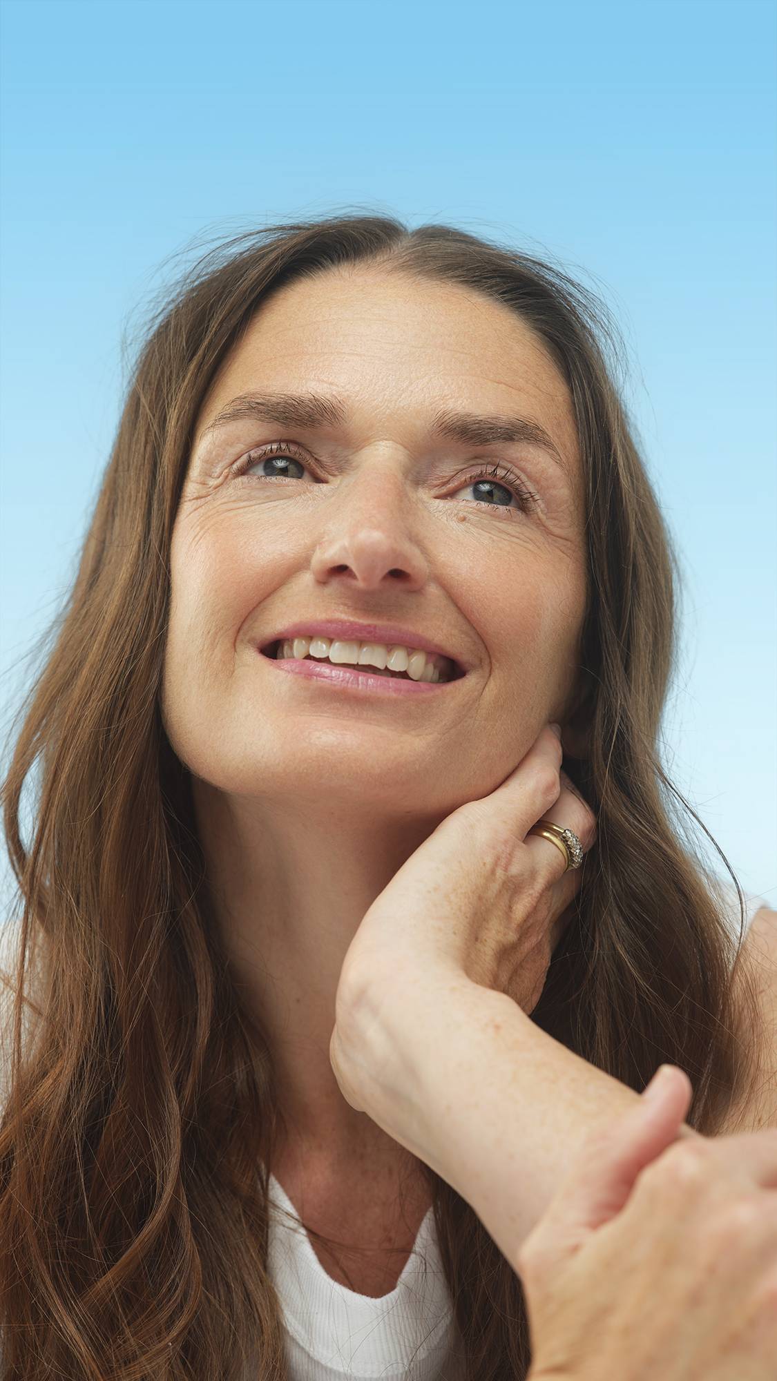 A close-up vertical portrait of a smiling person with medium, neutral-toned skin and long brown hair, looking up. They wear the Medium Neutral Beauty Balm and gently touch their neck, set against a bright blue background.