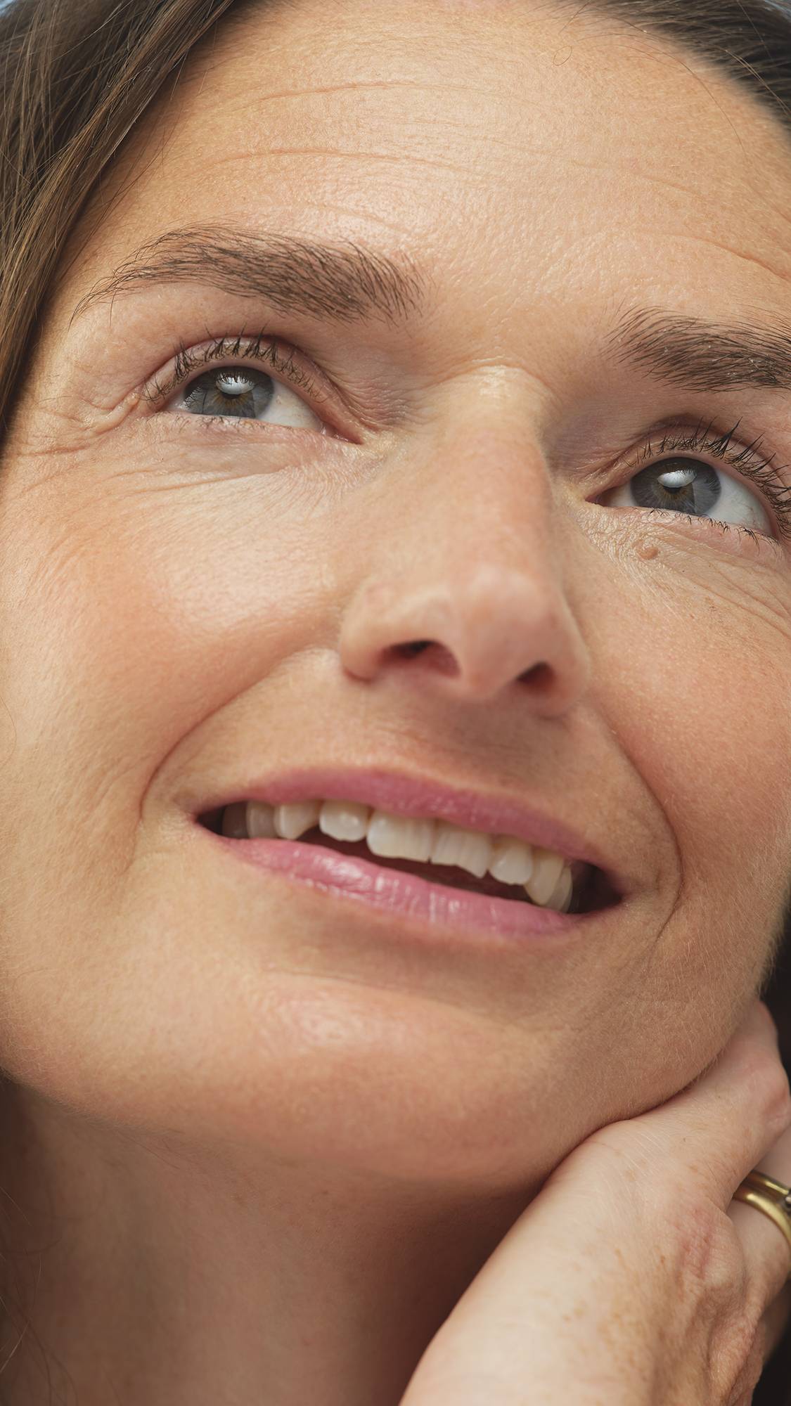 A close-up vertical shot of a smiling person's face, looking up, highlighting their eyes and mouth. The Medium Neutral Beauty Balm provides a smooth, natural-looking base on their medium skin.