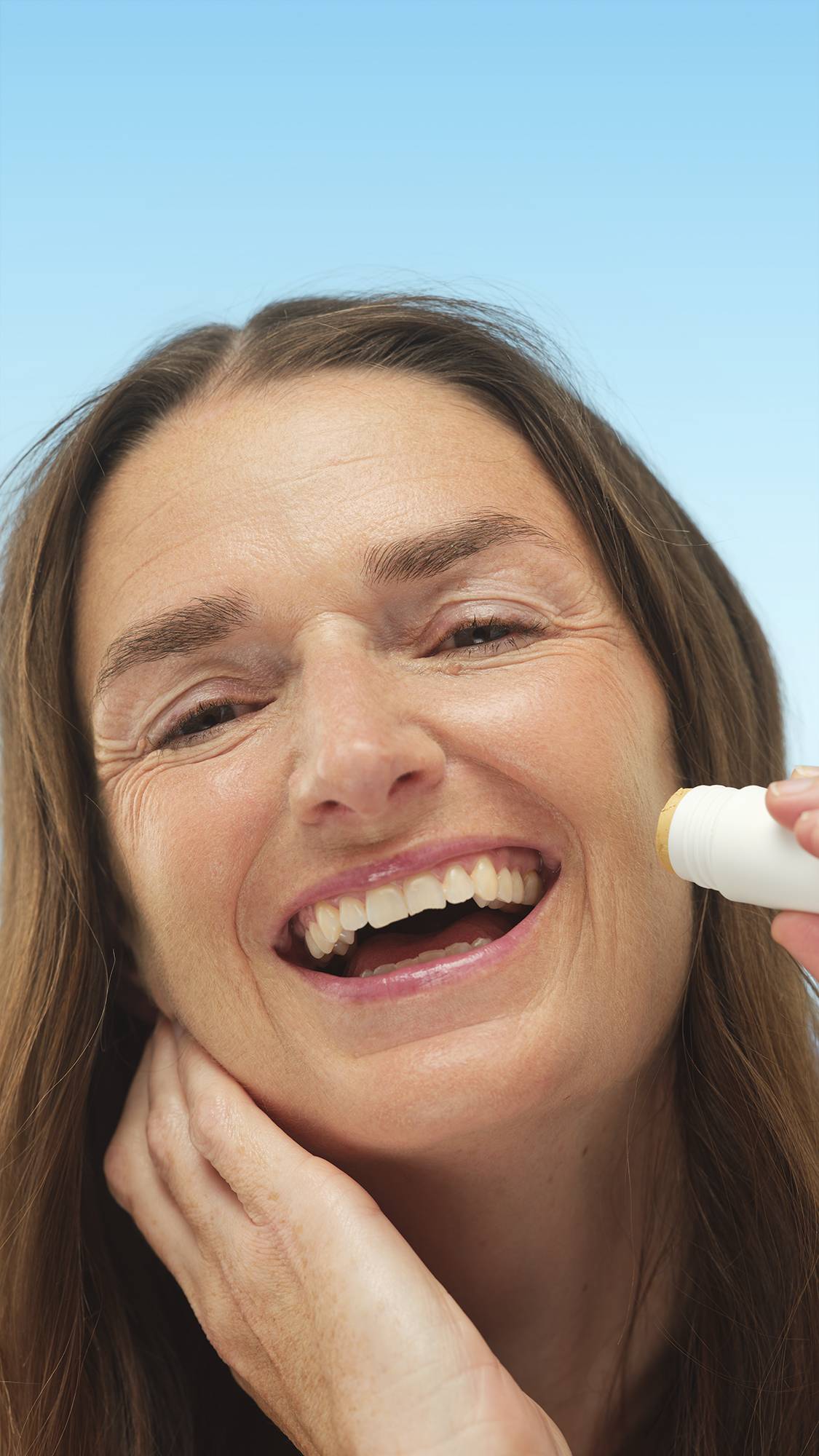 A close-up vertical portrait of a broadly smiling person with medium, neutral-toned skin, applying the white stick of Medium Neutral Beauty Balm directly to their cheek. They have long brown hair, set against a bright blue background.