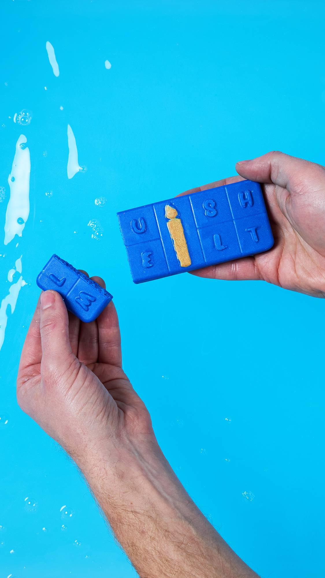 Two hands hold the Menorah Melt product over a bright blue bath or water surface. The main melt spells "LUSH" on top, while one segment spelling "M" has been broken off and is held separately. The golden candle feature is visible on the main piece.