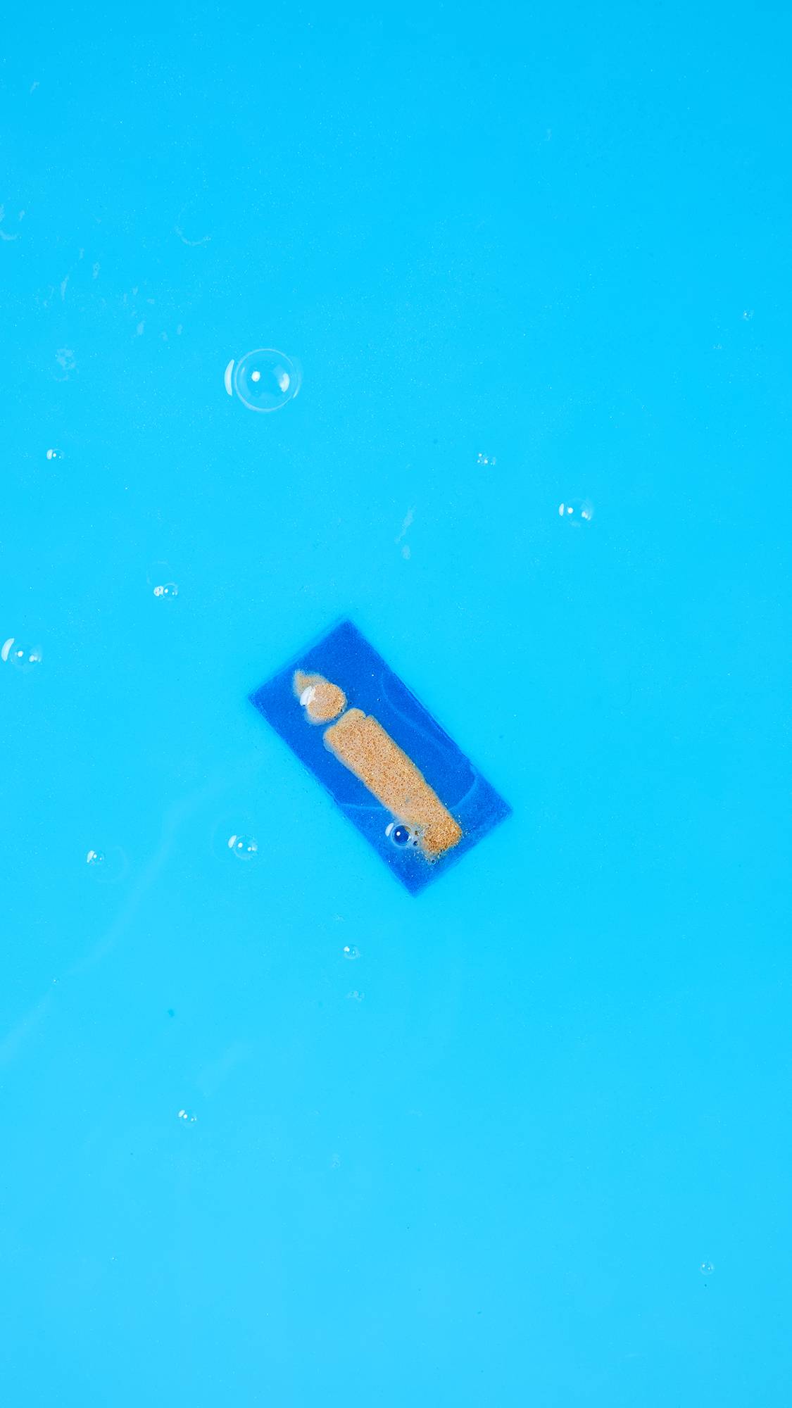 A single, rectangular blue segment of the Menorah Melt floats diagonally on the surface of bright blue water with a few bubbles. The golden orange candle or flame design is clearly visible on the floating piece.