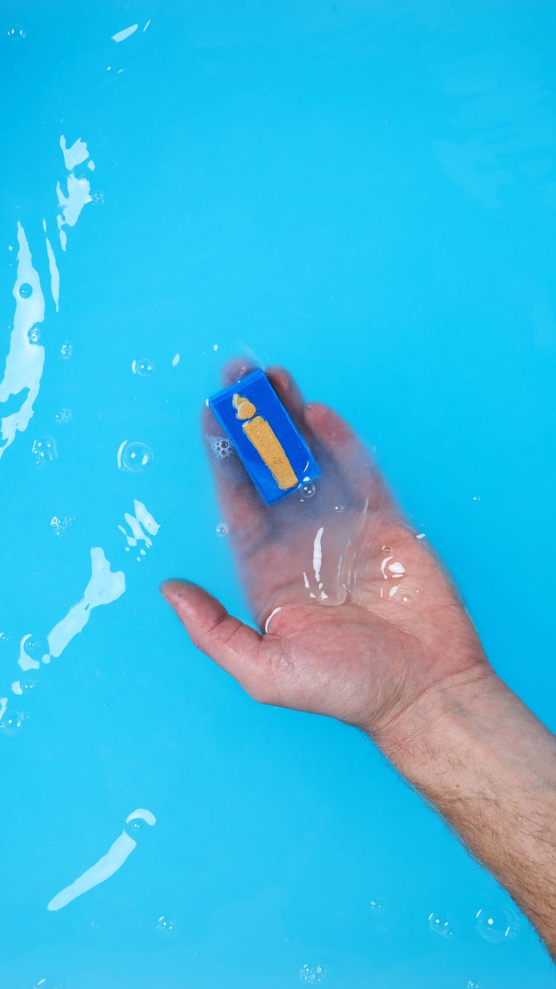 A hand holds a single, rectangular blue segment of the Menorah Melt just below the surface of the bright blue bath water. The golden orange candle or flame design is clearly visible on the piece.
