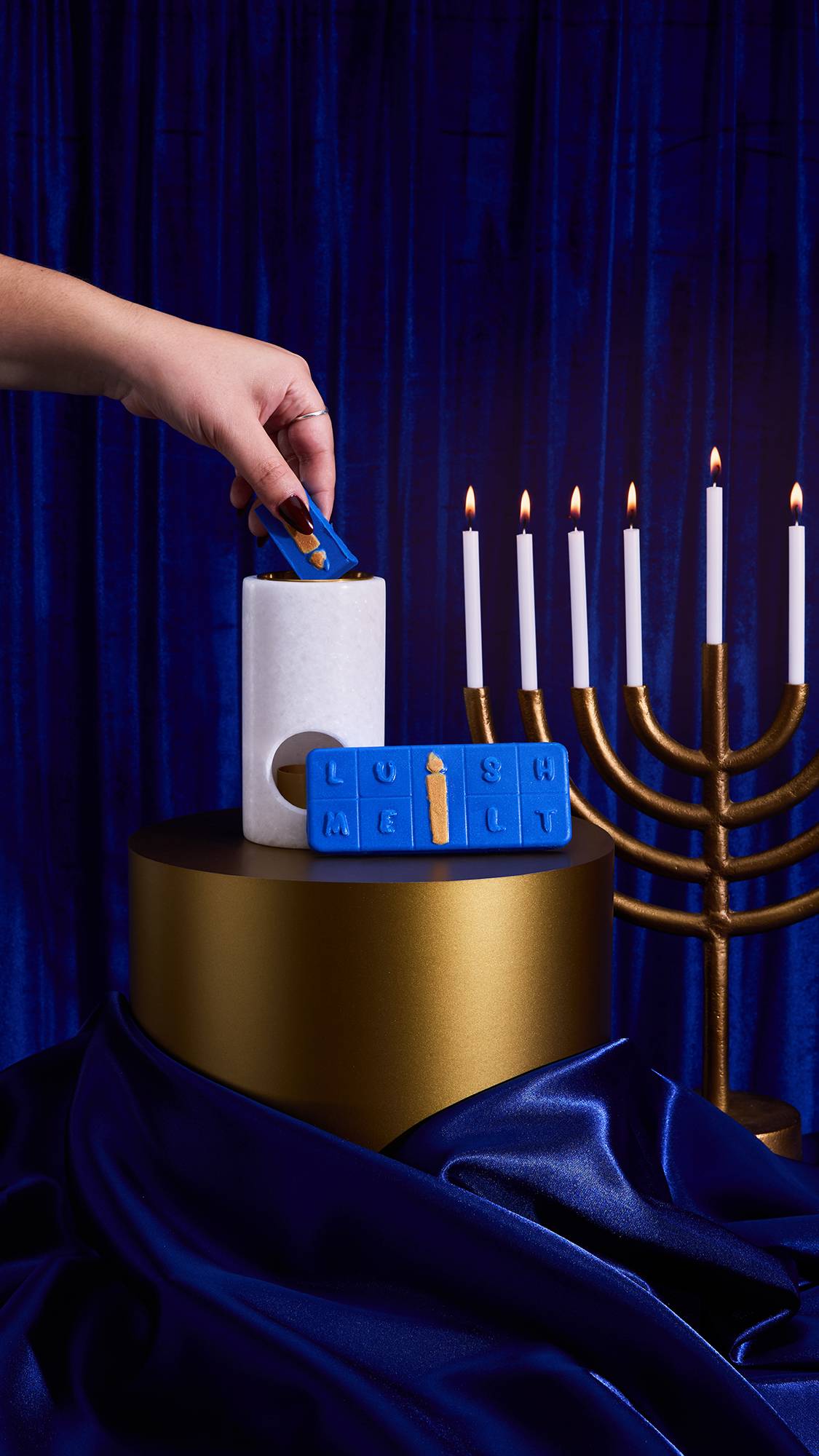 A dramatic scene features the Menorah Melt against royal blue velvet. A hand places a blue segment into a white warmer on a gold cylinder. The rest of the melt and a gold menorah with six lit candles are nearby.