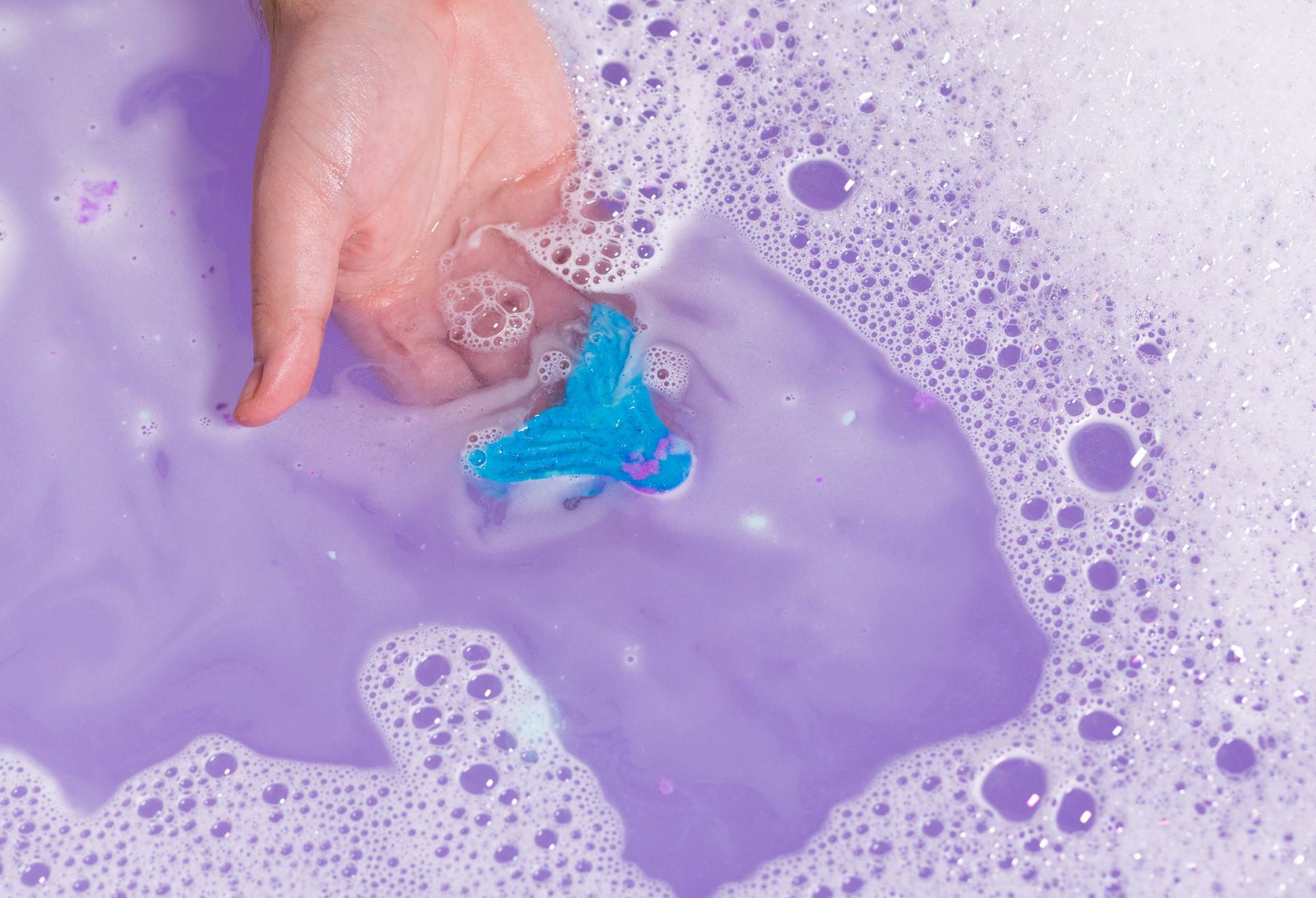 A hand holds the Mermaid Tail in purple water. All that is visible is the blue fin with bubbles forming on the surface.