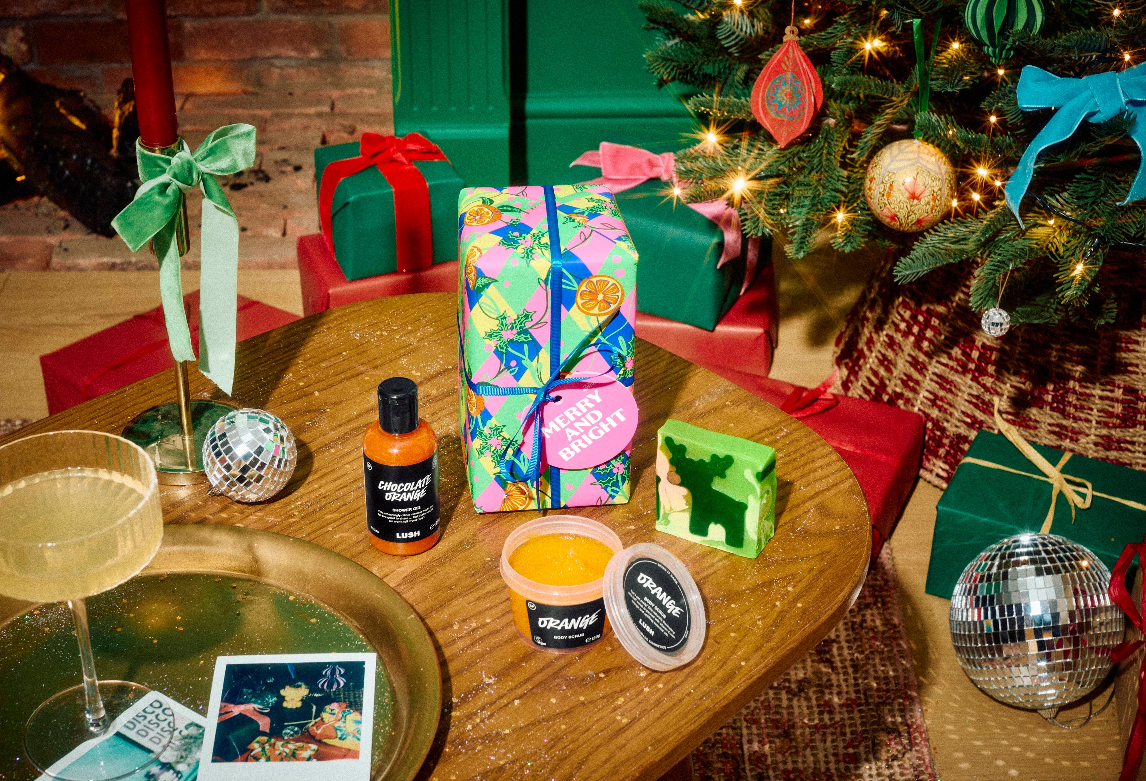 The Lush Merry and Bright gift set sits on a wooden table with Chocolate Orange shower gel, Orange body scrub, and a green reindeer soap, surrounded by wrapped presents and a decorated Christmas tree.