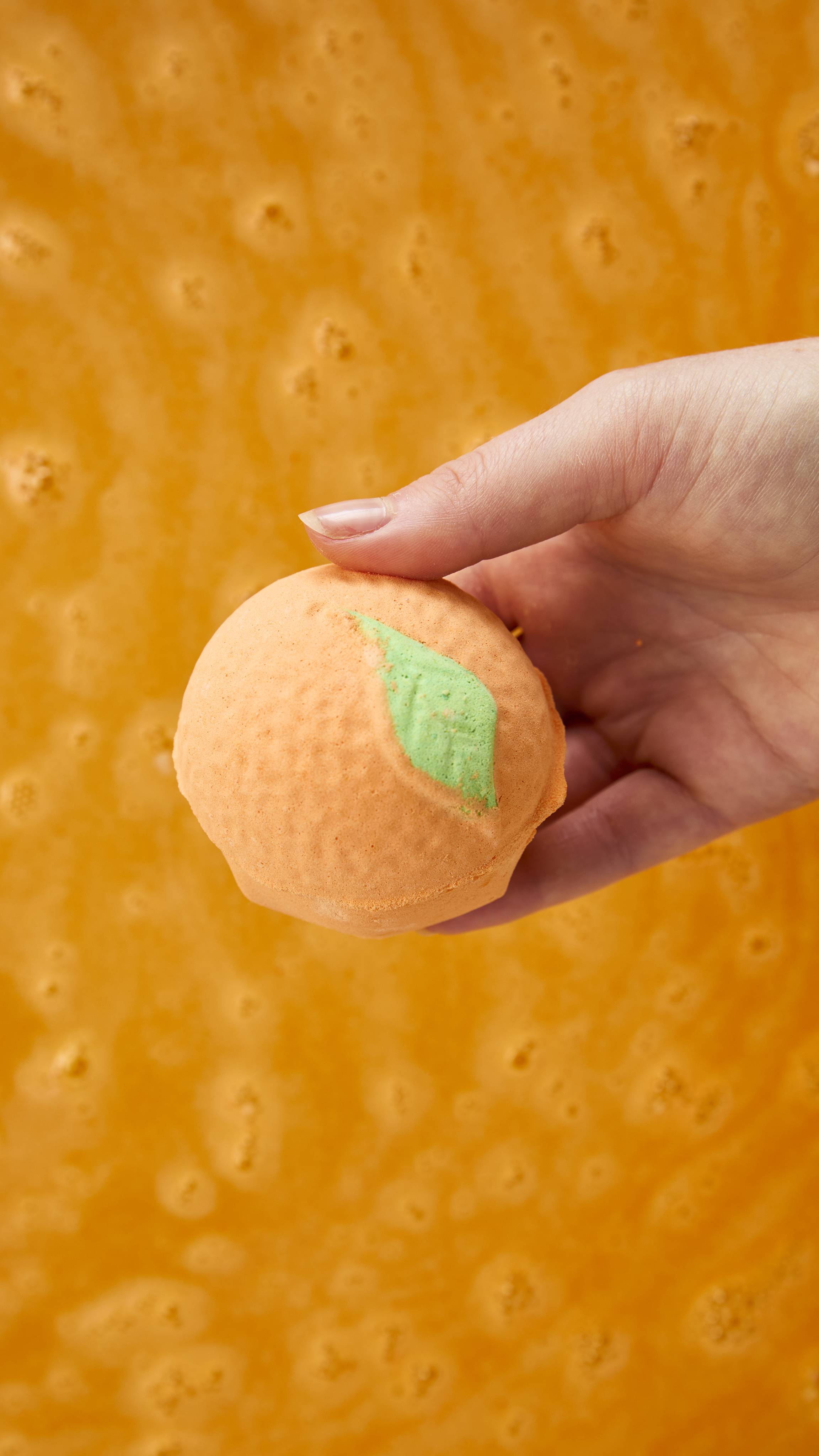 A hand holds a round, orange-shaped bath bomb featuring a green leaf detail, positioned over swirling, frothy orange bathwater.