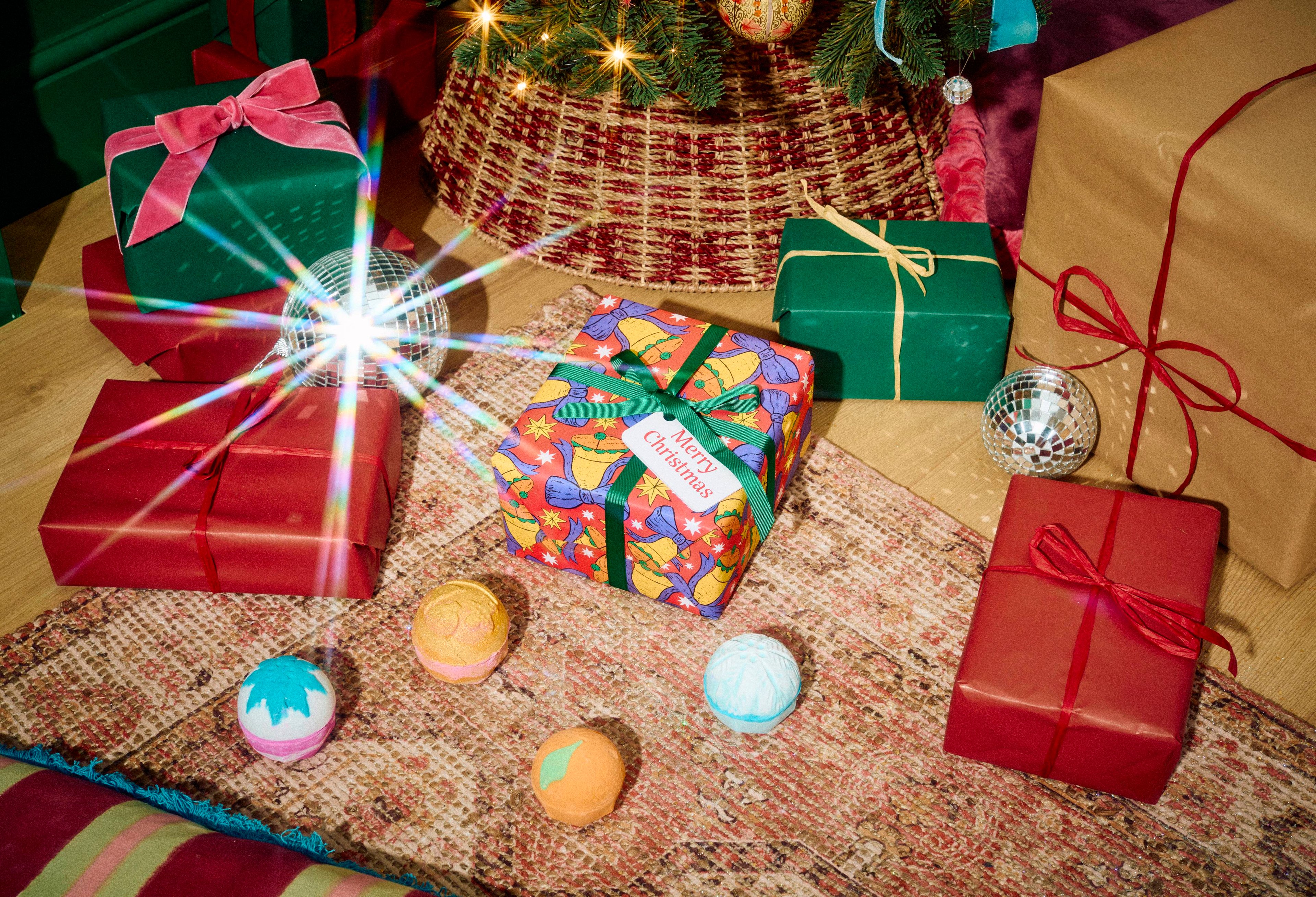 The Lush Merry Christmas gift set sits on a patterned rug surrounded by four colourful bath bombs, positioned in a festive room filled with wrapped presents beneath a decorated Christmas tree.