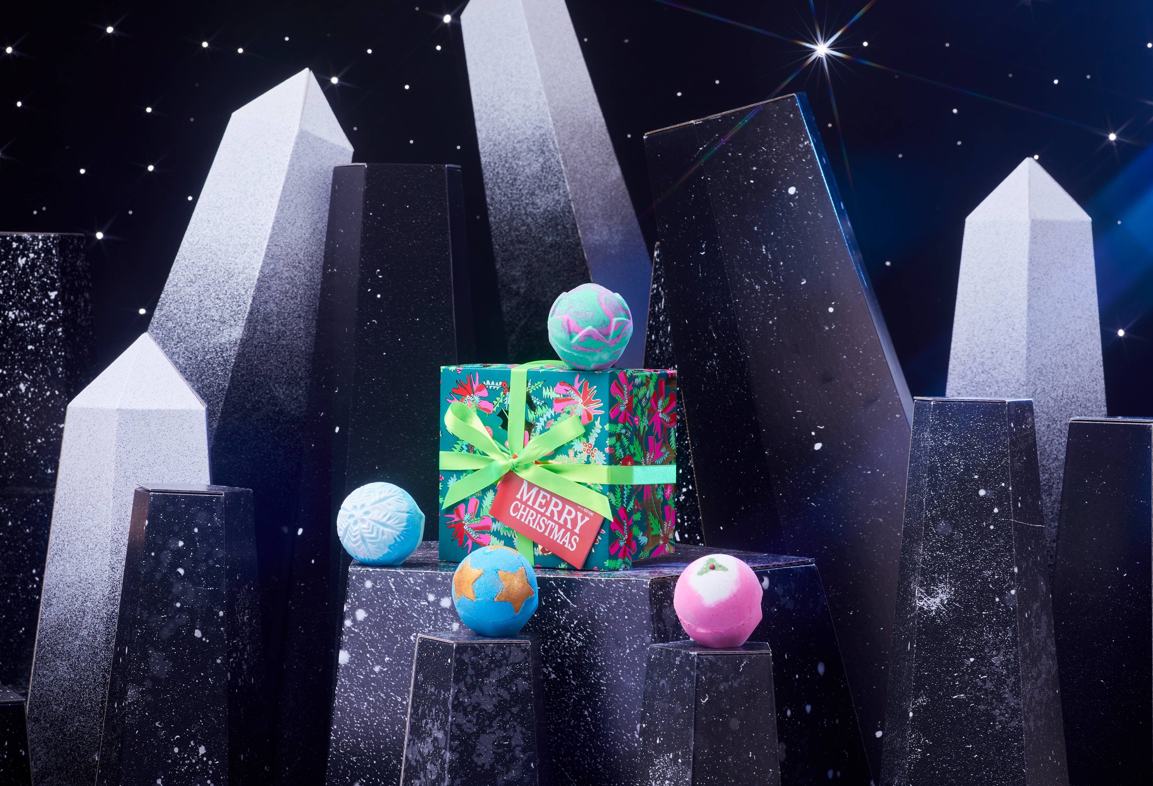 Merry Christmas gift box and 4 Christmas bath bombs are on a dark chromatic platform among geometric shapes. 