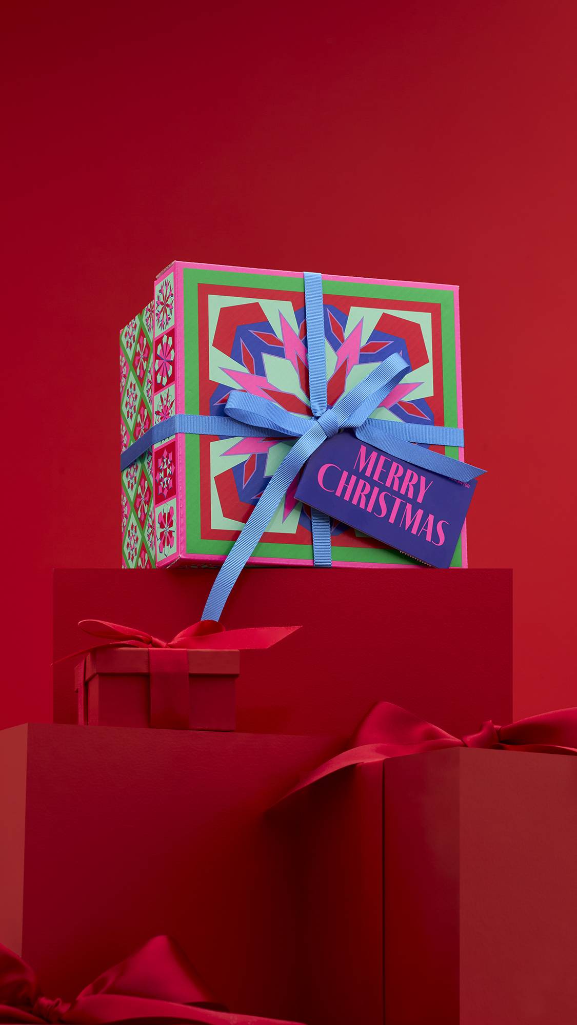 The image shows the colourful Merry Christmas gift box, tied with a blue ribbon and a deep purple gift tag, sitting on a solid red platform against a matching red background. 