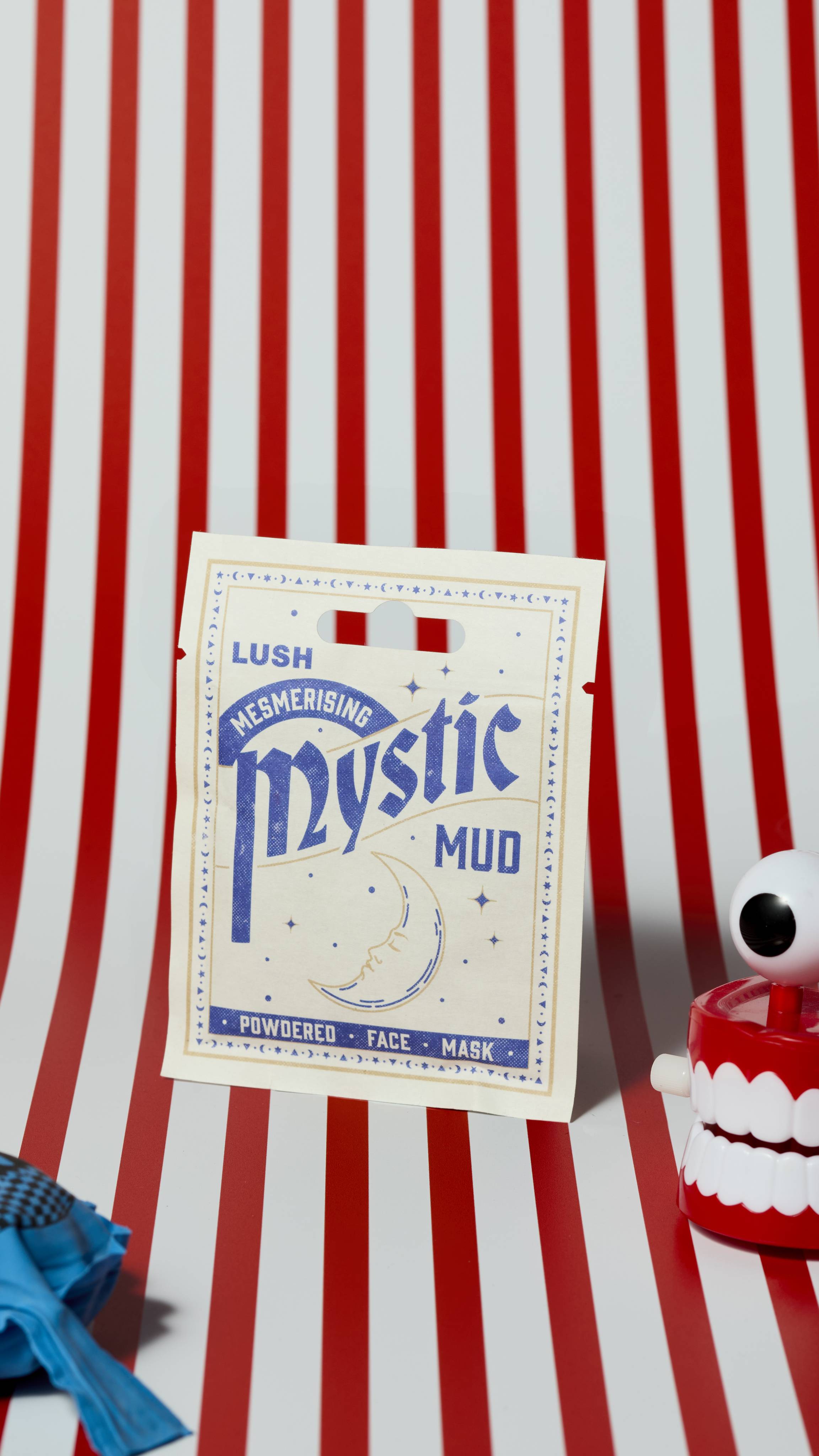 The image shows the Mystic Mud powdered face mask product sachet on a red and white, vertically striped background. There is a whoopie cushion and wind-up chattering teeth that are just visible.