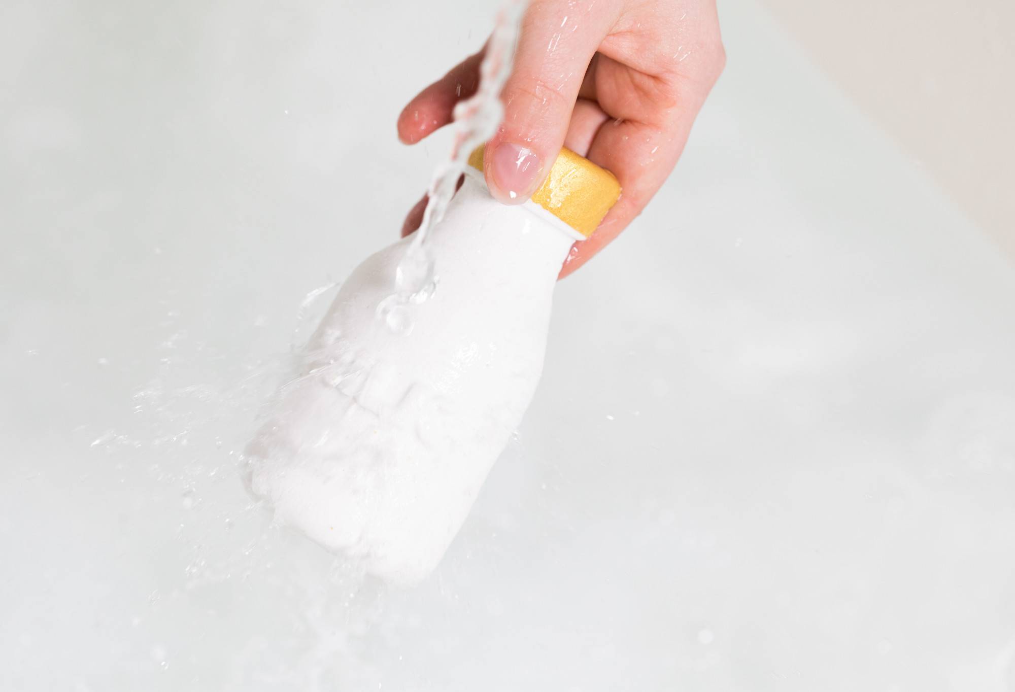 A hand holds a white Milky Bar by the gold lid as water runs over the bubble bar. In the background creamy water forms bubbles.