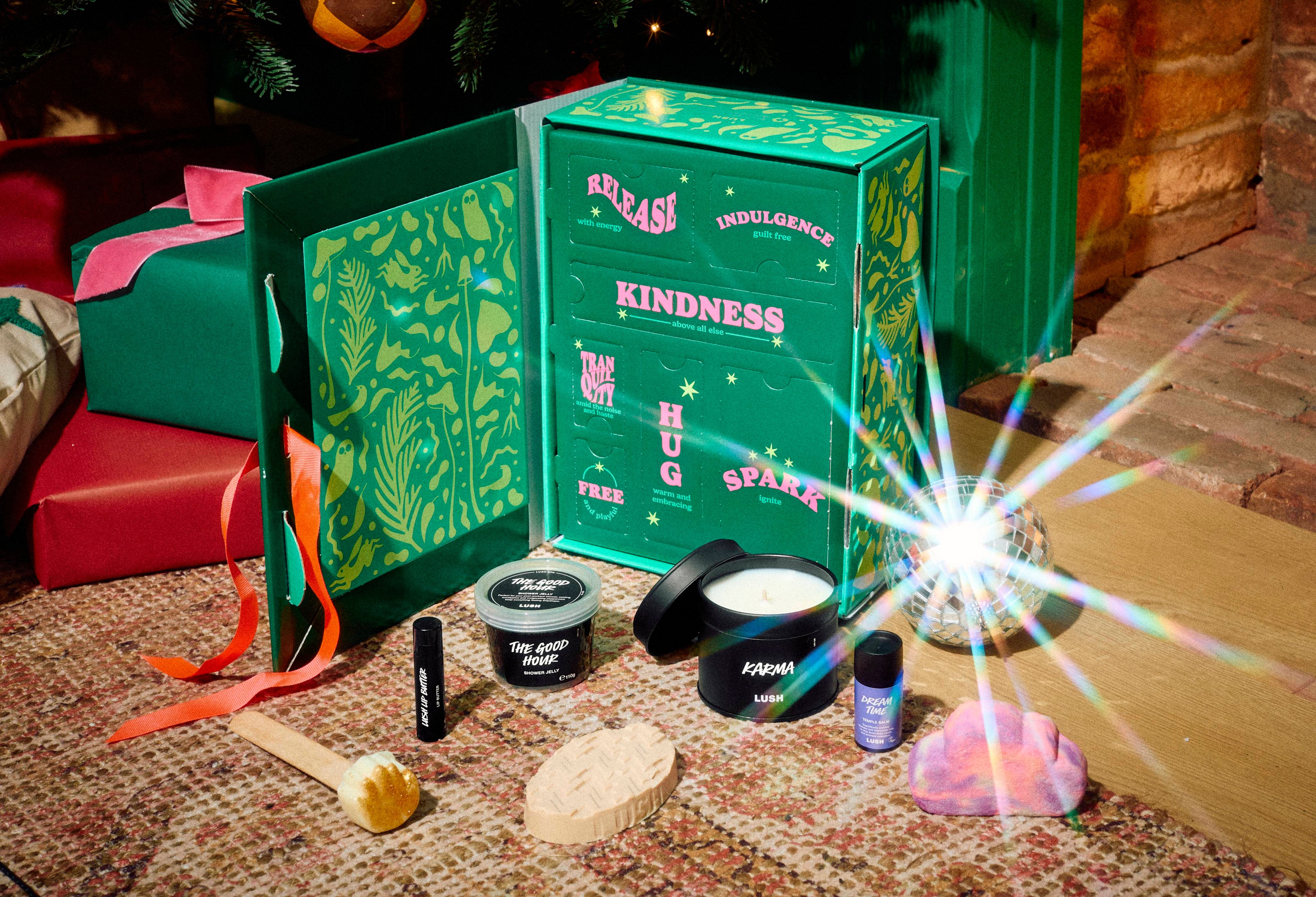 An open green Lush gift box with patterned doors displaying words like 'Kindness'. Spread out in front are bath and body products, including a candle, a solid hand mask on a stick, lip balm, and a small disco ball reflecting light.
