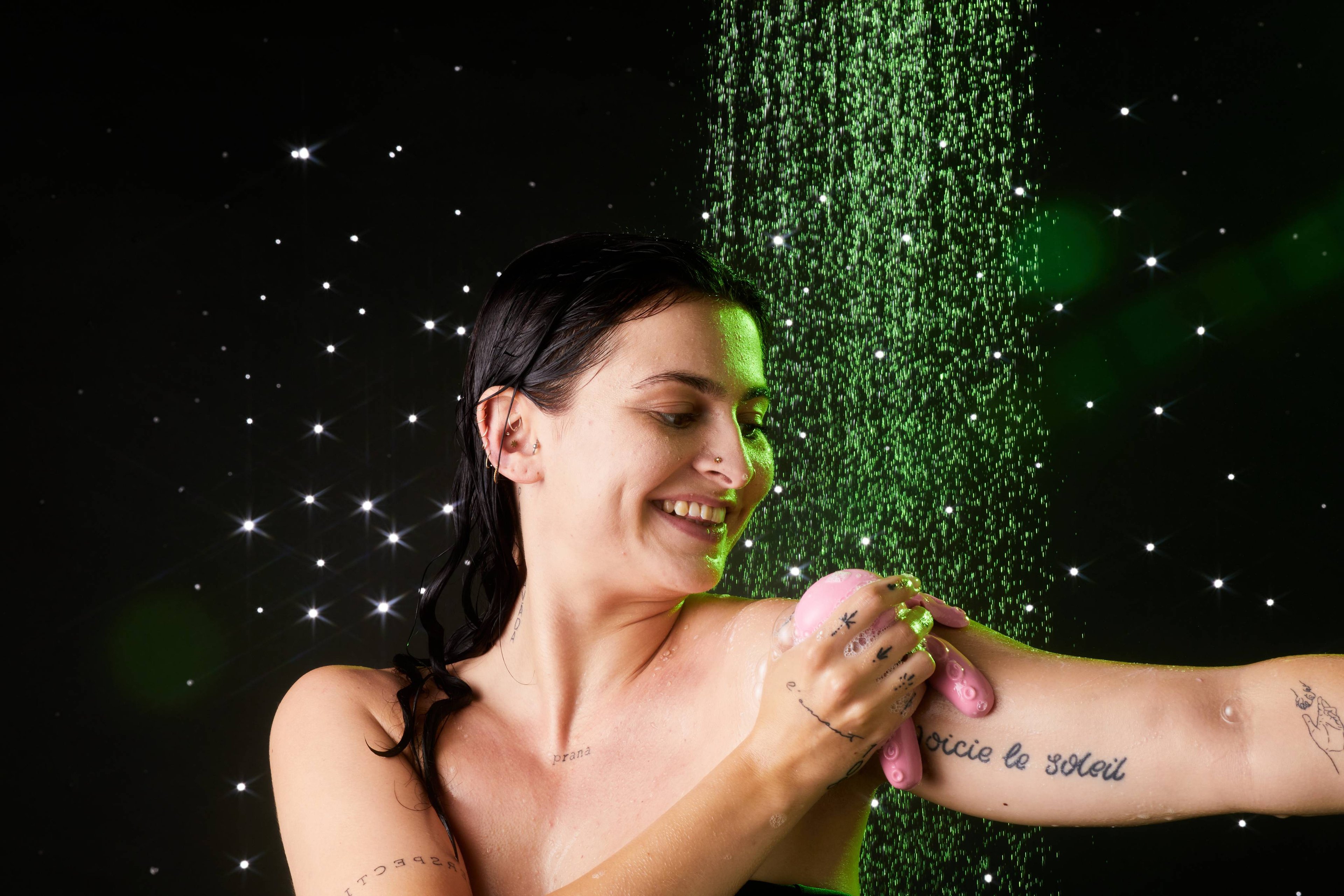 Model is under shower water as they smooth the tentacled, shower jelly over their shoulder and arm on a twinkling background.