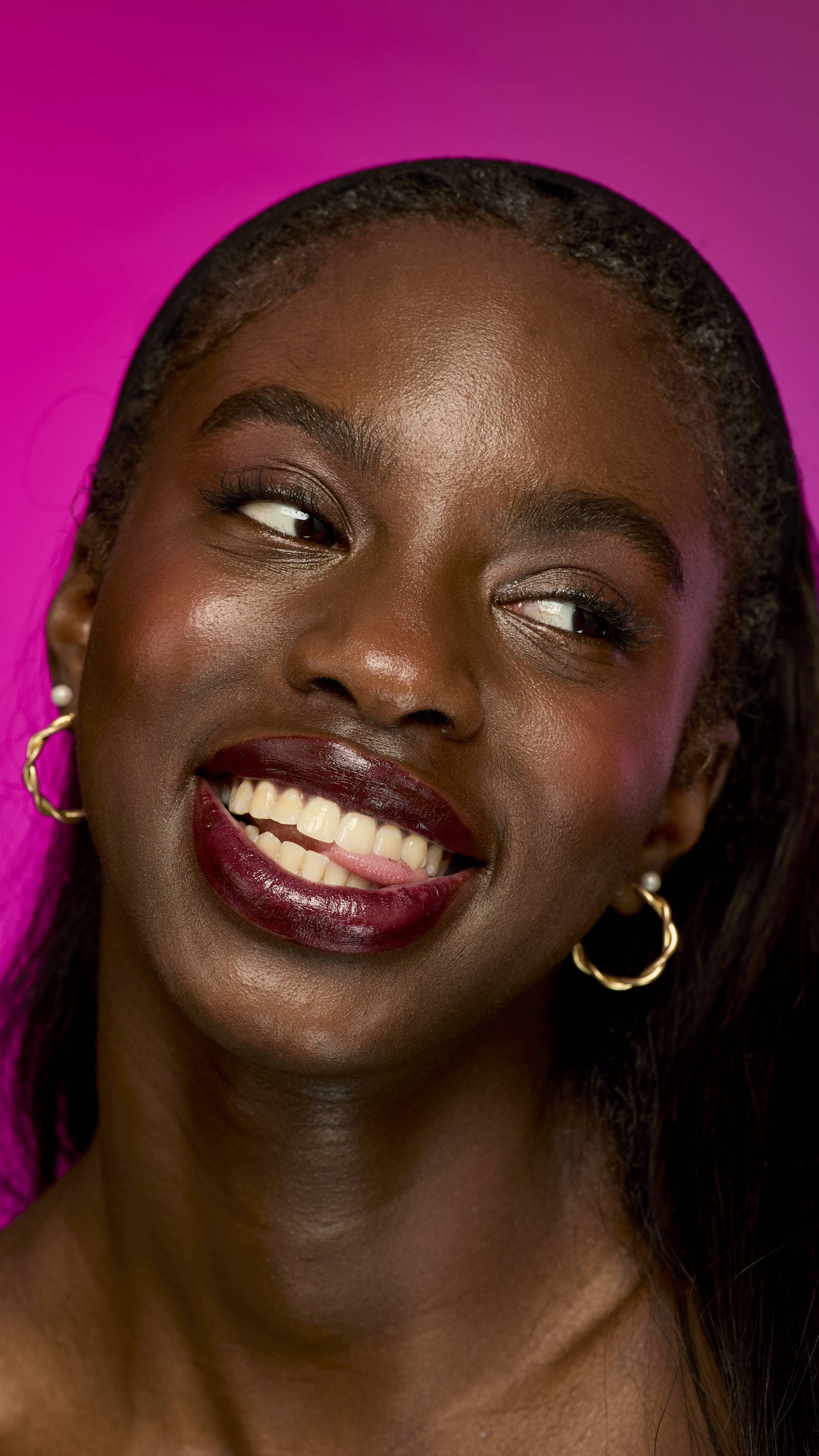 A smiling person with deep skin tone and long, straight dark hair is shown from the neck up on a vibrant magenta background. They are wearing the velvety, deep red wine-coloured Moonshiner lipstick.
