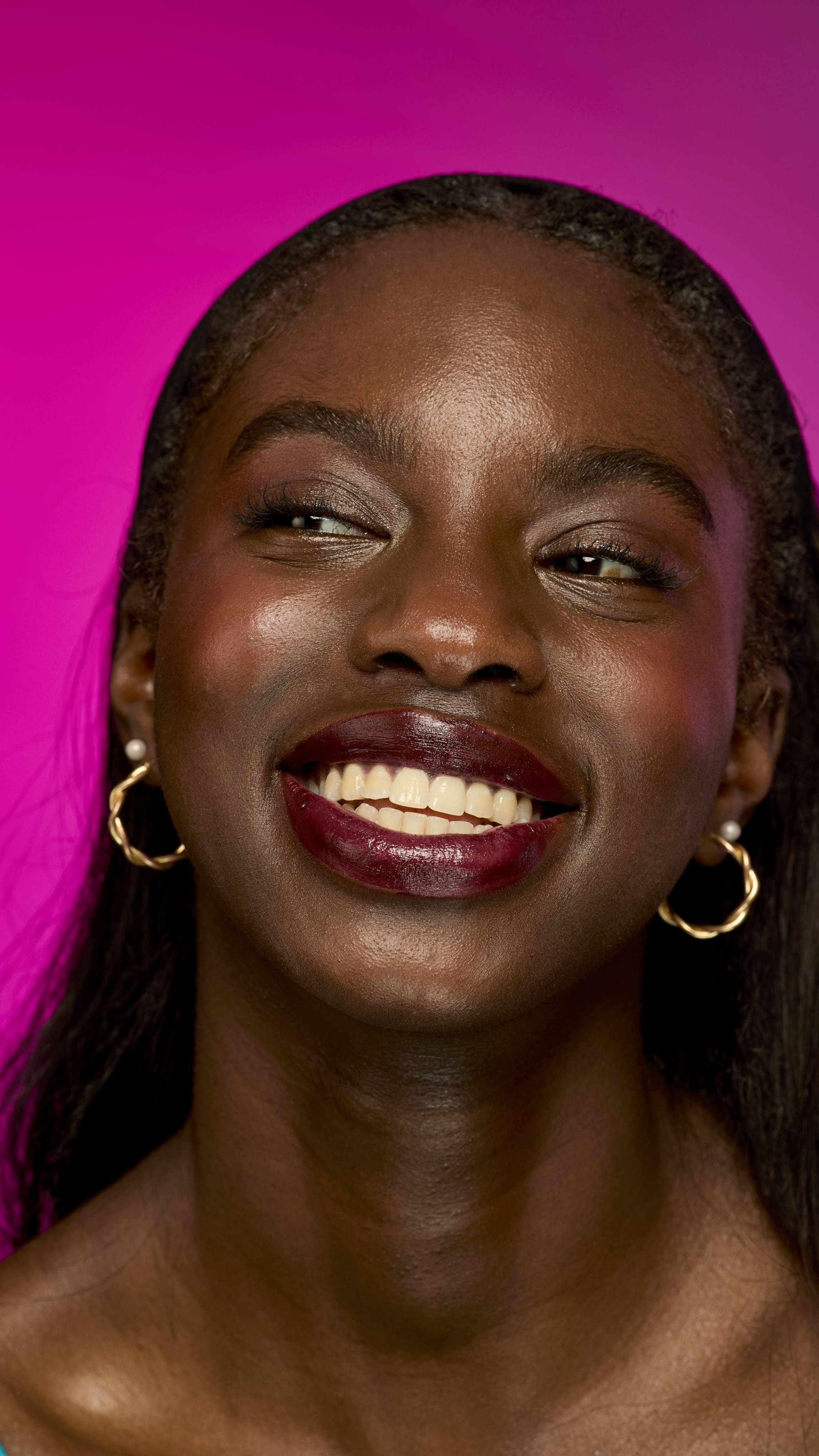 A smiling person with deep skin tone and long, straight dark hair is shown from the neck up on a vibrant magenta background. They are wearing the velvety, deep red wine-coloured Moonshiner lipstick.