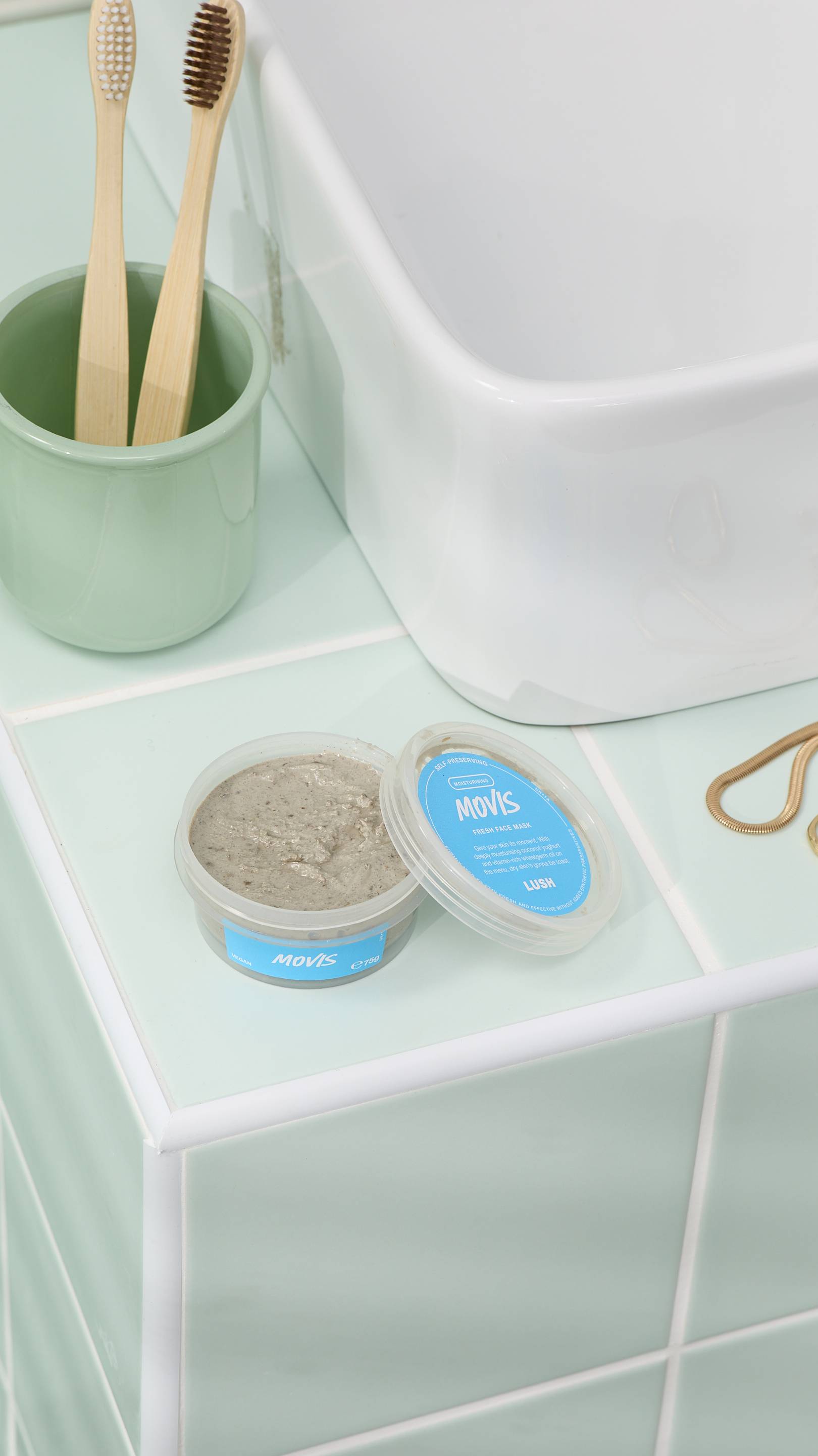 The image shows an open pot of grey, textured "Movis" face mask sitting on light green bathroom tiles next to a white sink. In the background, two bamboo toothbrushes are in a green cup. Gold jewellery rests nearby on the tiled surface.