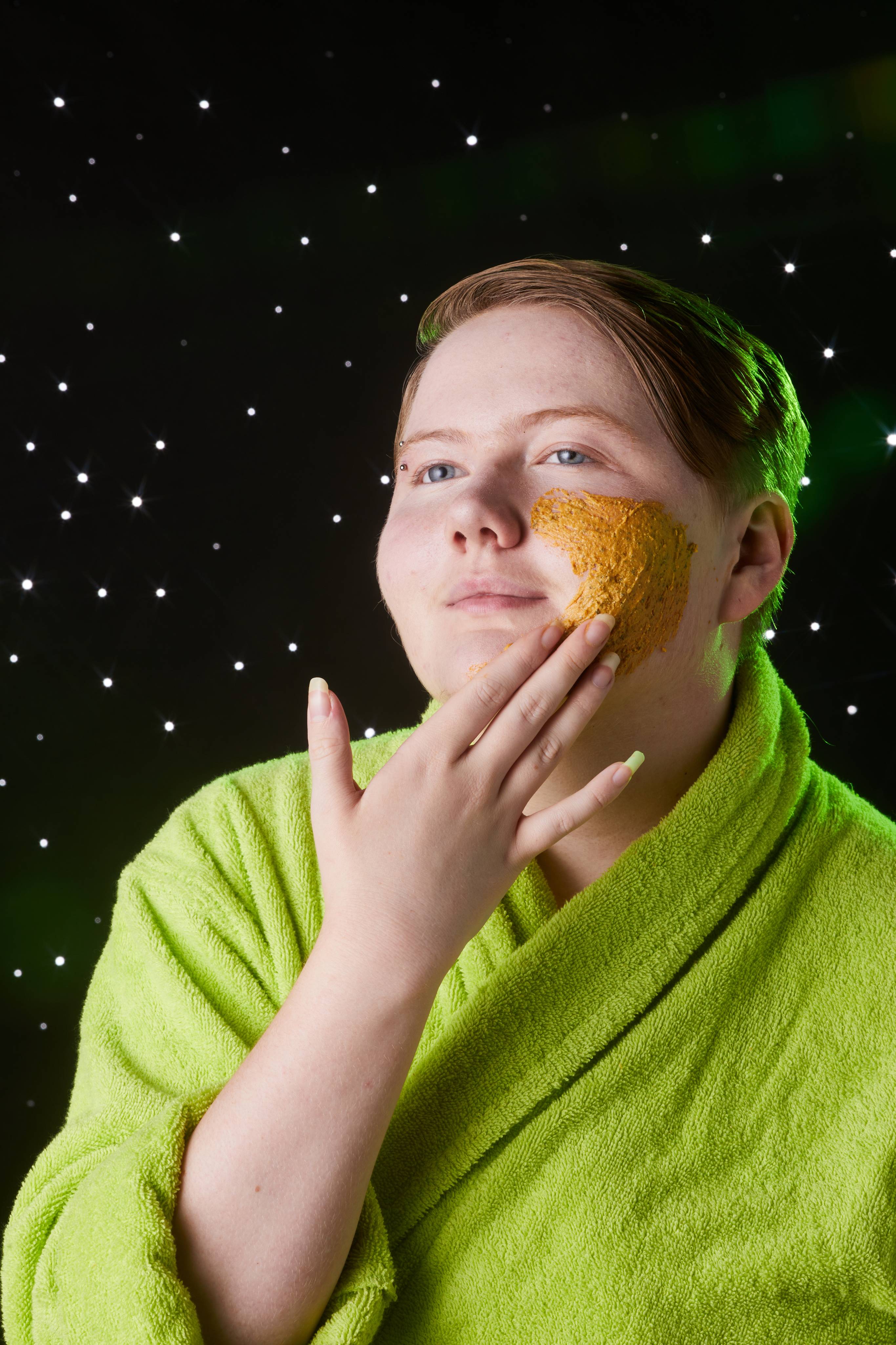 Image shows model in a neon-green robe as they gently spread the product over their face with their hand.