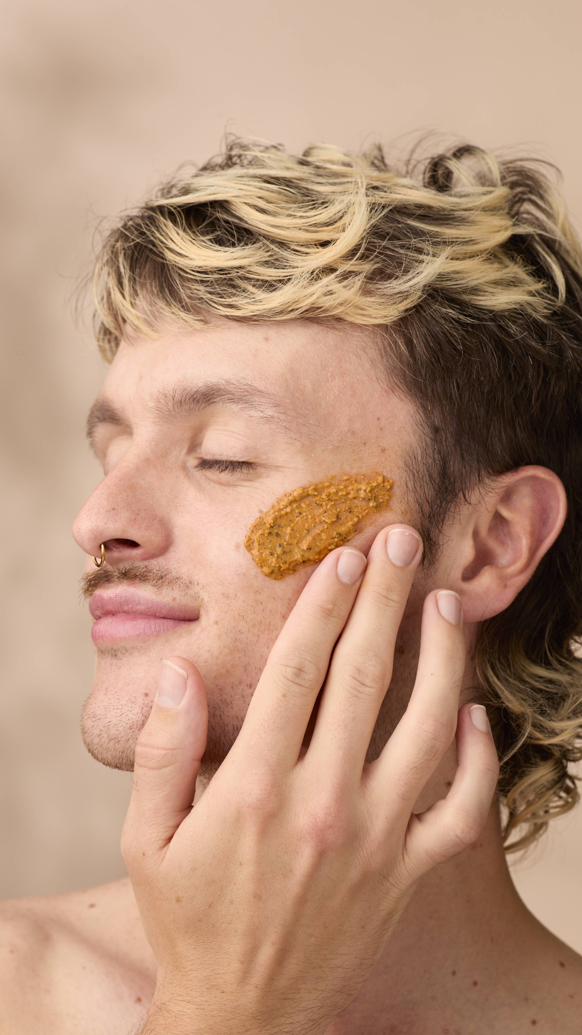 A close-up profile view of a person with blonde-highlighted hair and eyes closed, gently applying Lush Mr Pumpkin Face fresh face mask onto their cheek with two fingers. They have a nose ring and a slight smile against a neutral backdrop.